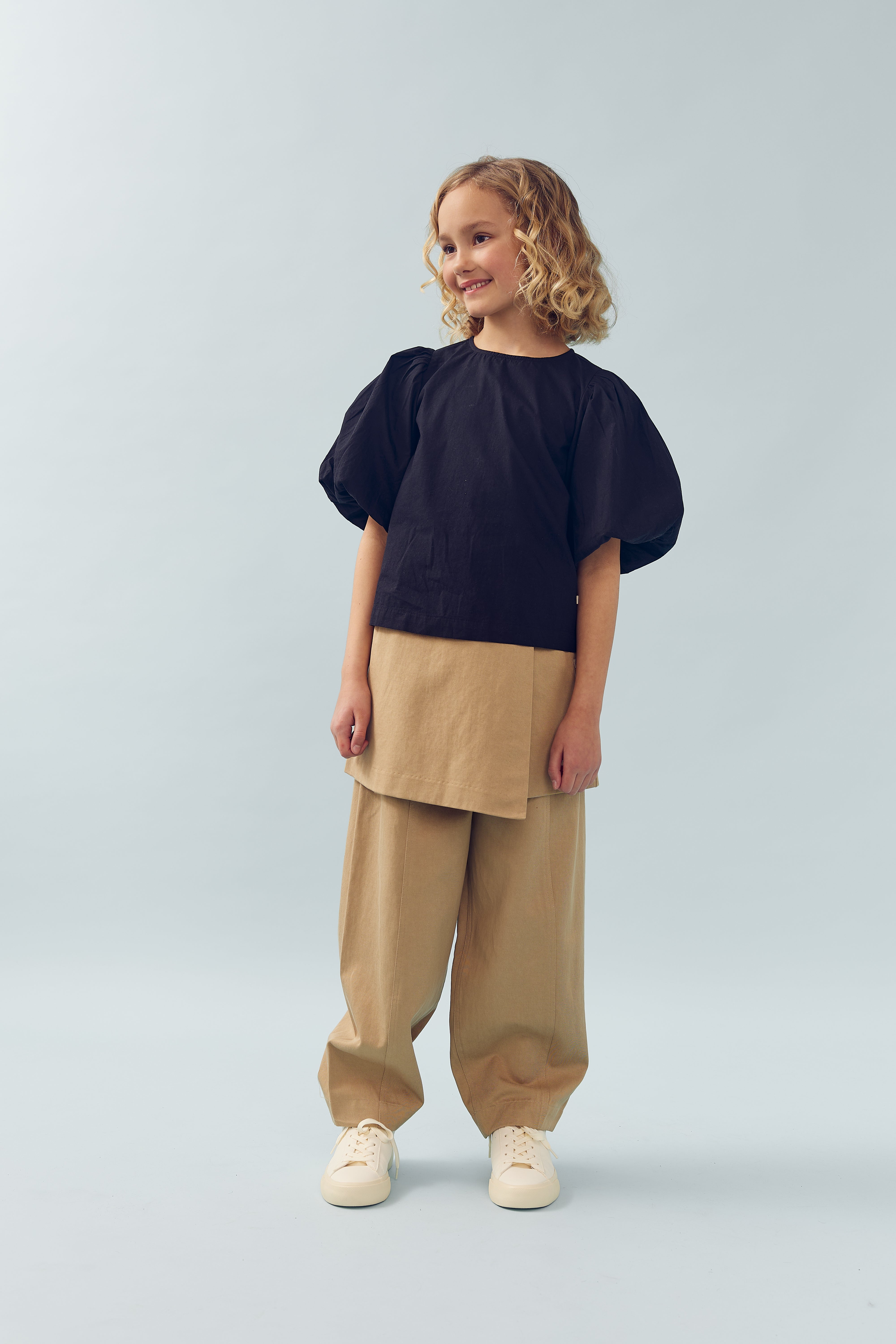 Kind draagt een zwarte blouse met wijde mouwen en beige broek, lachend in een lichte, minimalistische achtergrond.