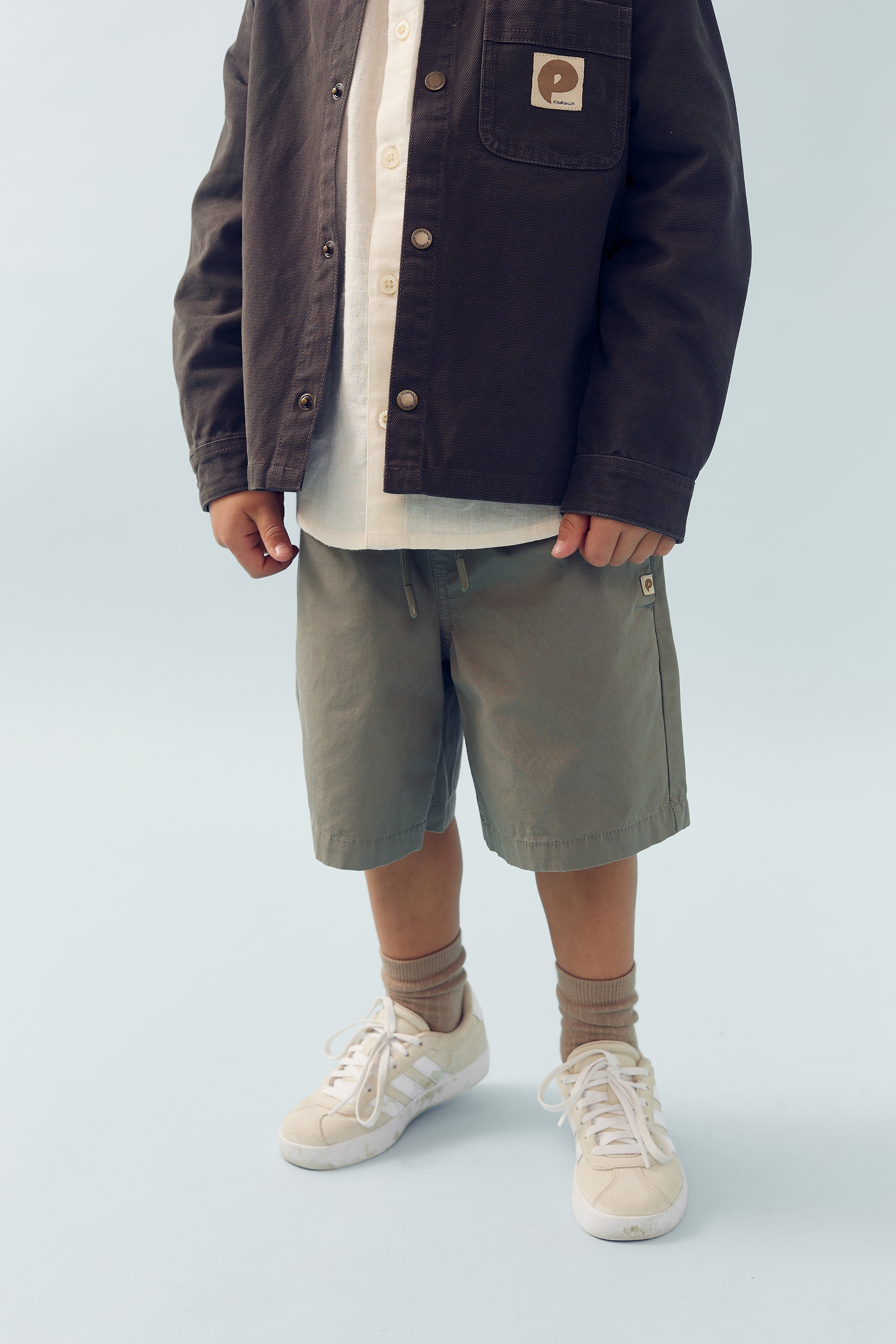 Een kind draagt een bruine jas, licht shirt en grijze korte broek, met beige sokken en sneakers. Op de jas zit een vierkante patch met een gebogen logo. De achtergrond is lichtblauw.