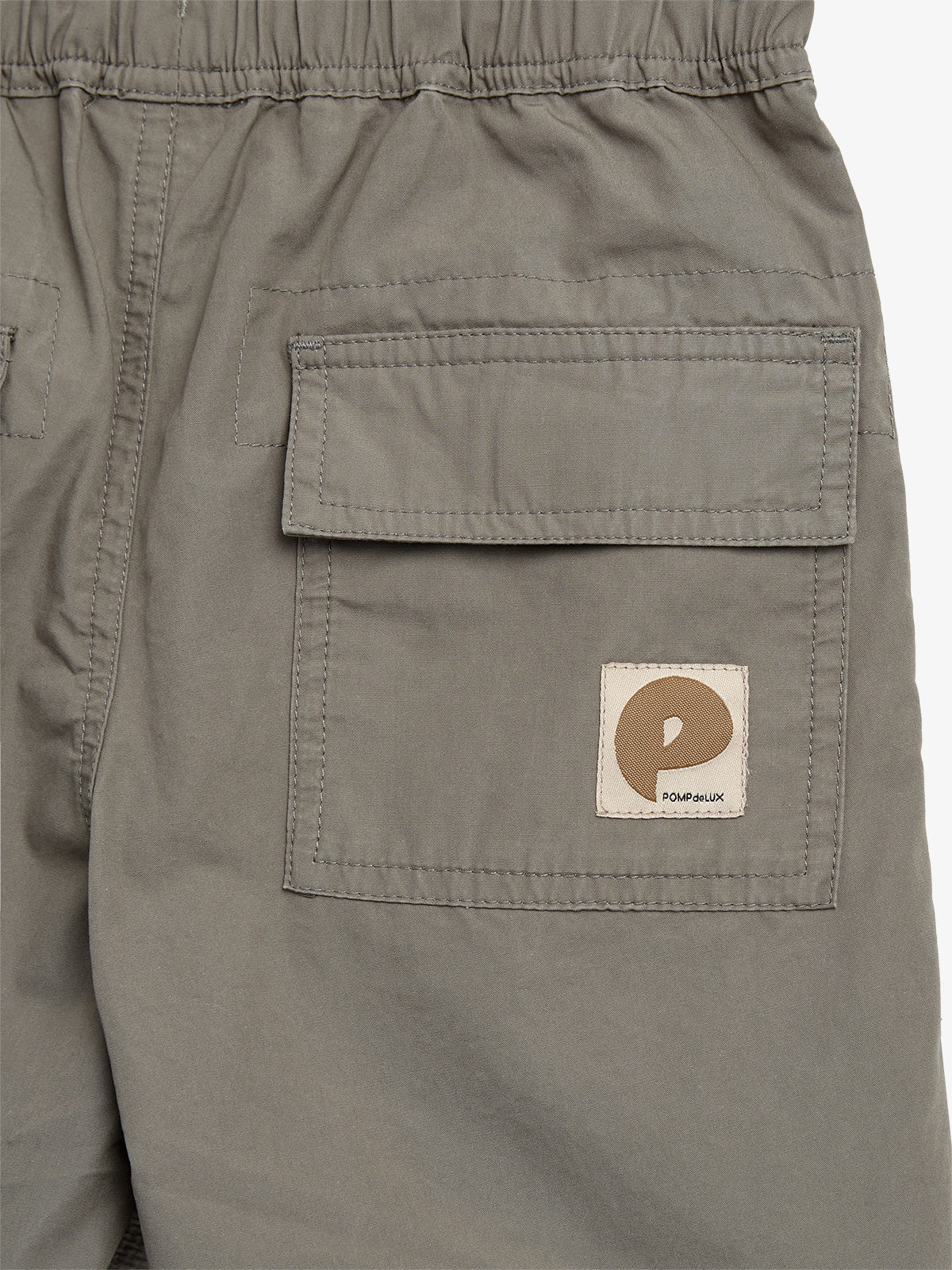Een grijze broekzak met klep, heeft een logo patch met een bruine "P" en de tekst "POMPdeLUX." De achtergrond is egaal van kleur.