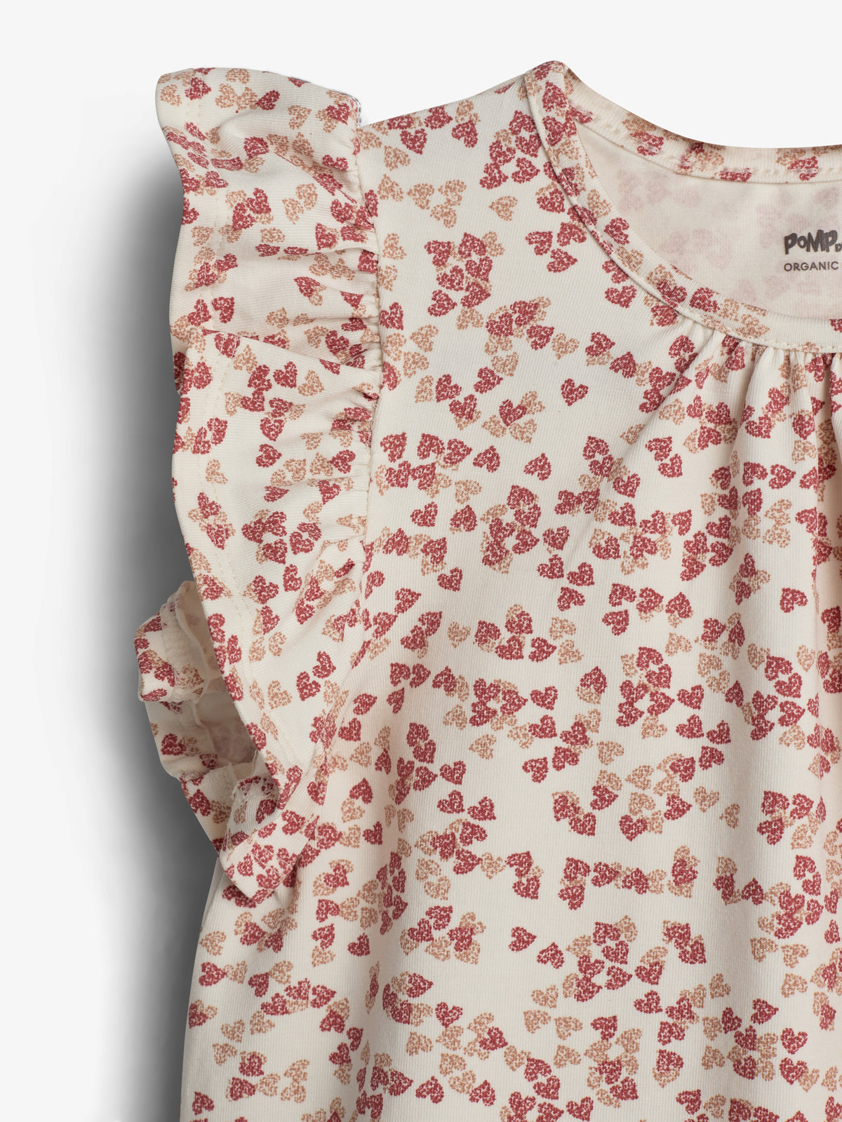 Een crèmekleurig meisjes t-shirt met rode en roze bloemmotieven, heeft ruches bij de mouwen. Het label binnenin vermeldt "POMPdeLUX ORGANIC COTTON," aangeduid op een gladde achtergrond.