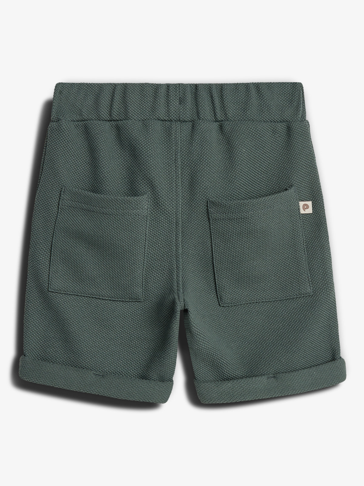 Een groene, structuurstof korte broek met opgerolde pijpen, heeft twee grote achterzakken en een elastische tailleband. De achtergrond is eenvoudig en grijs. Rechts zit een klein label.