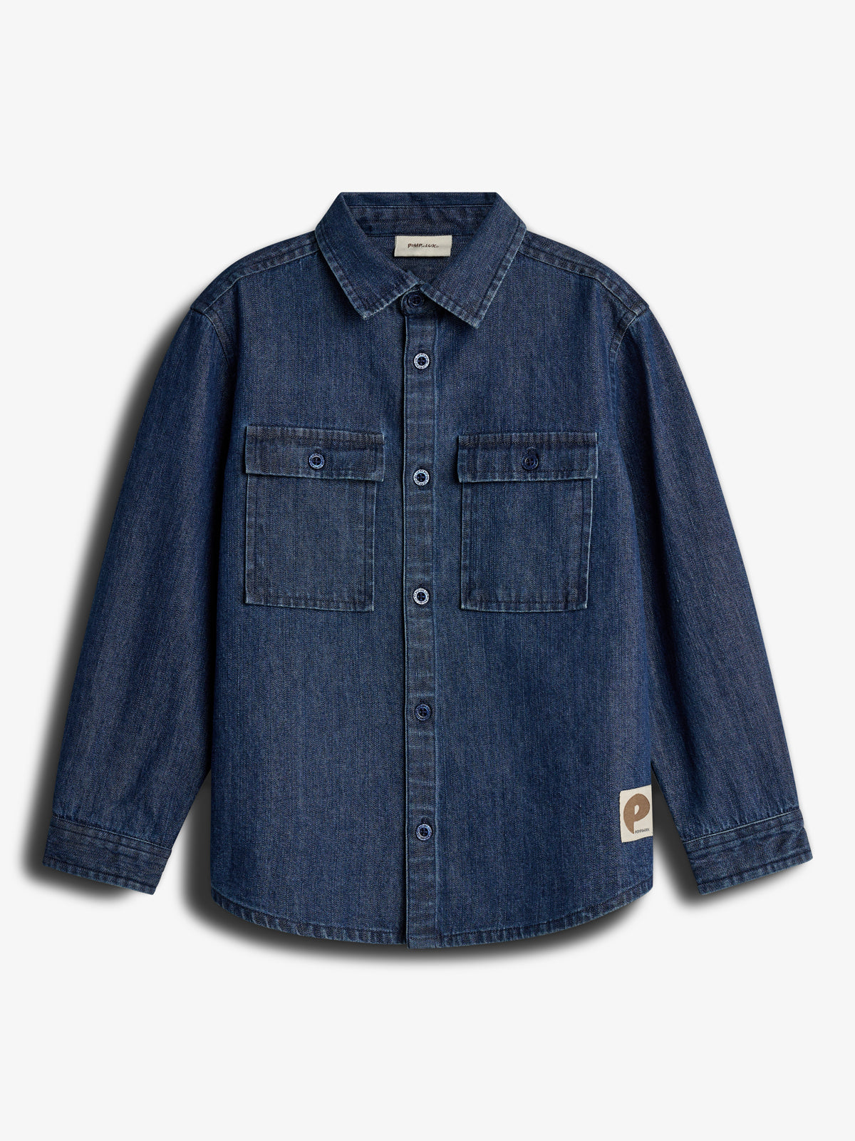 Een denim overhemd hangt los, met knoopsluiting en twee borstzakken, in een eenvoudige witte achtergrond. Er bevindt zich een kleine bruine label met een "P" aan de onderkant.