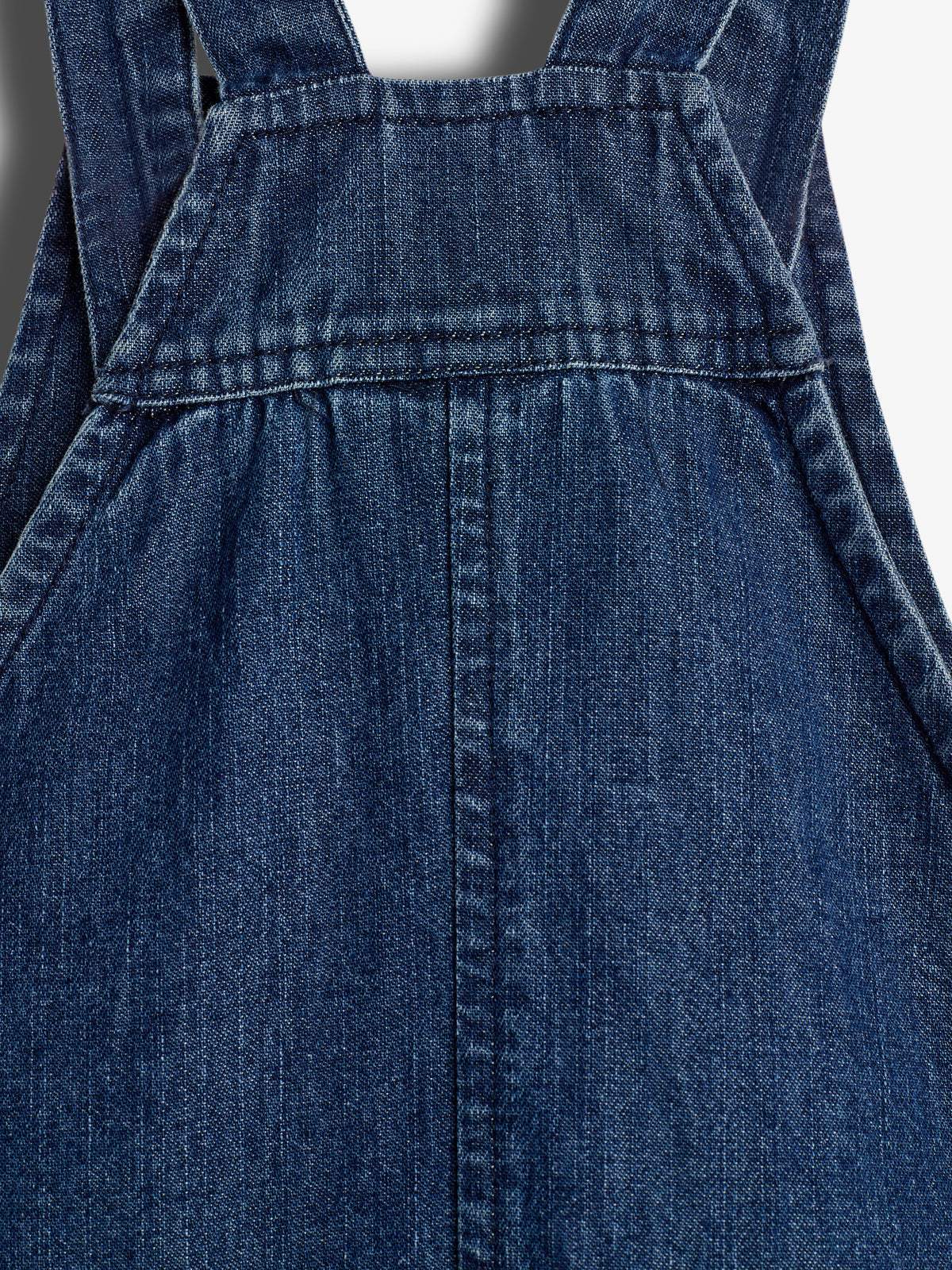 Denimstof vormt een schort met riemen aan de bovenkant. De kenmerkende stiknaden voegen detail toe, terwijl de rechte, donkere textuur de algehele uitstraling in een neutrale omgeving accentueert.