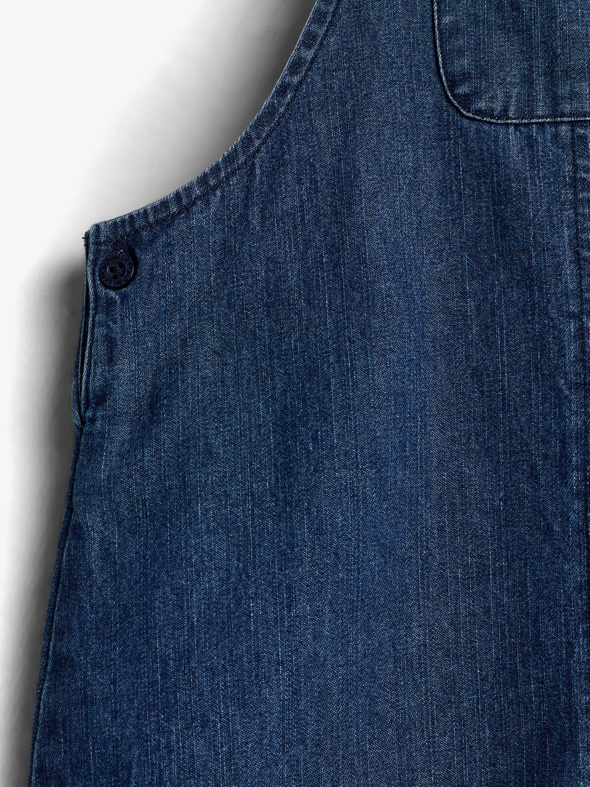 Een blauw denim hemd heeft een zichtbare naad en een knoop. Het hangt stil tegen een effen, lichtgekleurde achtergrond.