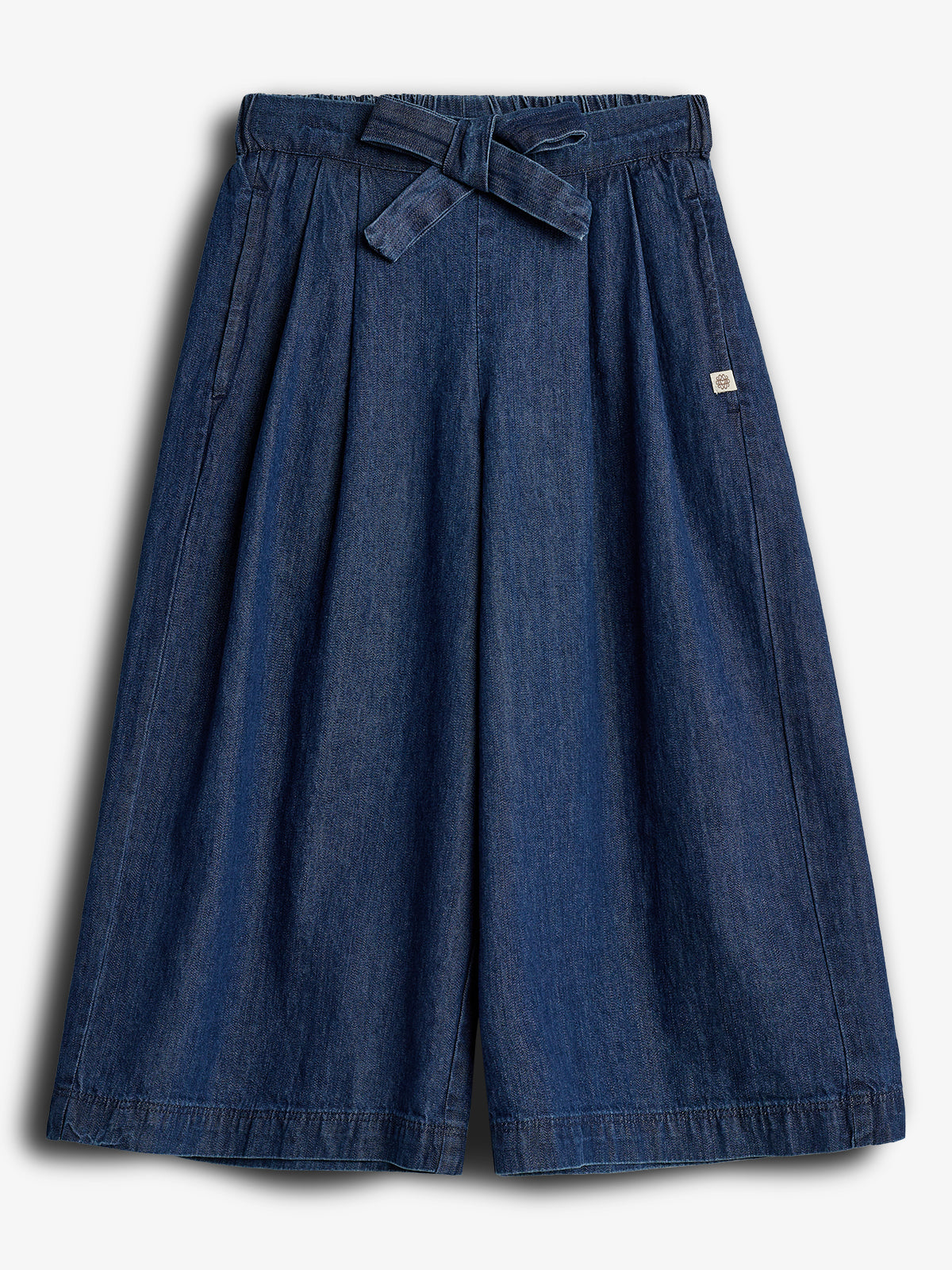 Een blauwe denim culotte met een strikriem en elastische tailleband, symmetrisch gelegen in een neutrale, witte achtergrond. Er is een klein, vierkant label aan de rechterkant.
