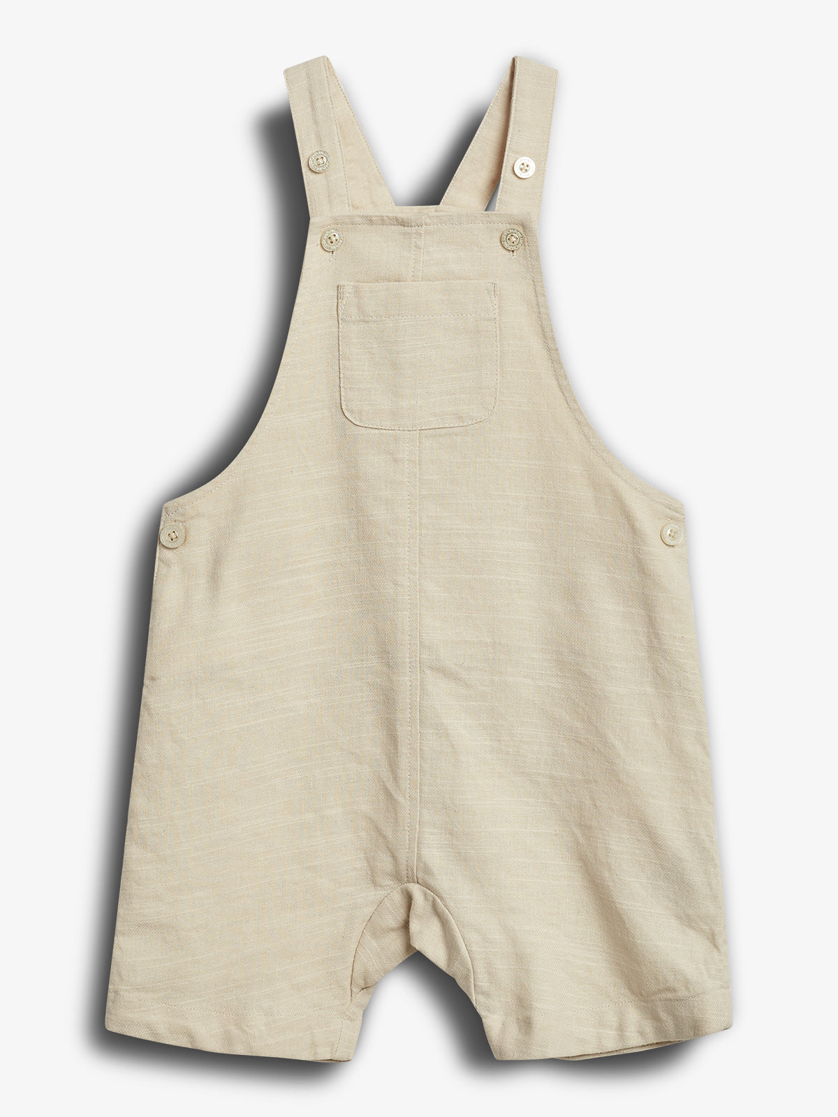 Een beige baby tuinbroek met knopen op de banden en zijkanten, een borstzak, hangt tegen een neutraal witte achtergrond.