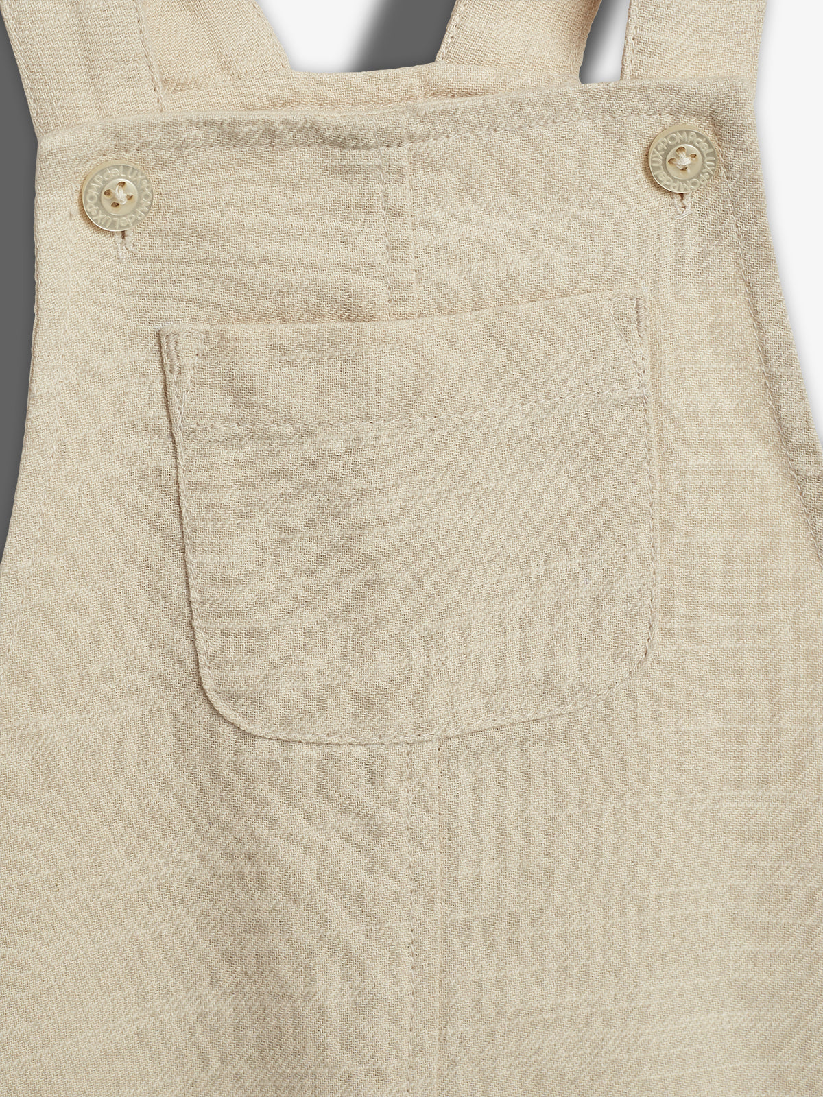 Beige linnen schort met knopen en een vierkante zak, gefotografeerd tegen een grijze achtergrond. Knopen bevatten de tekst "LOVEMADE".