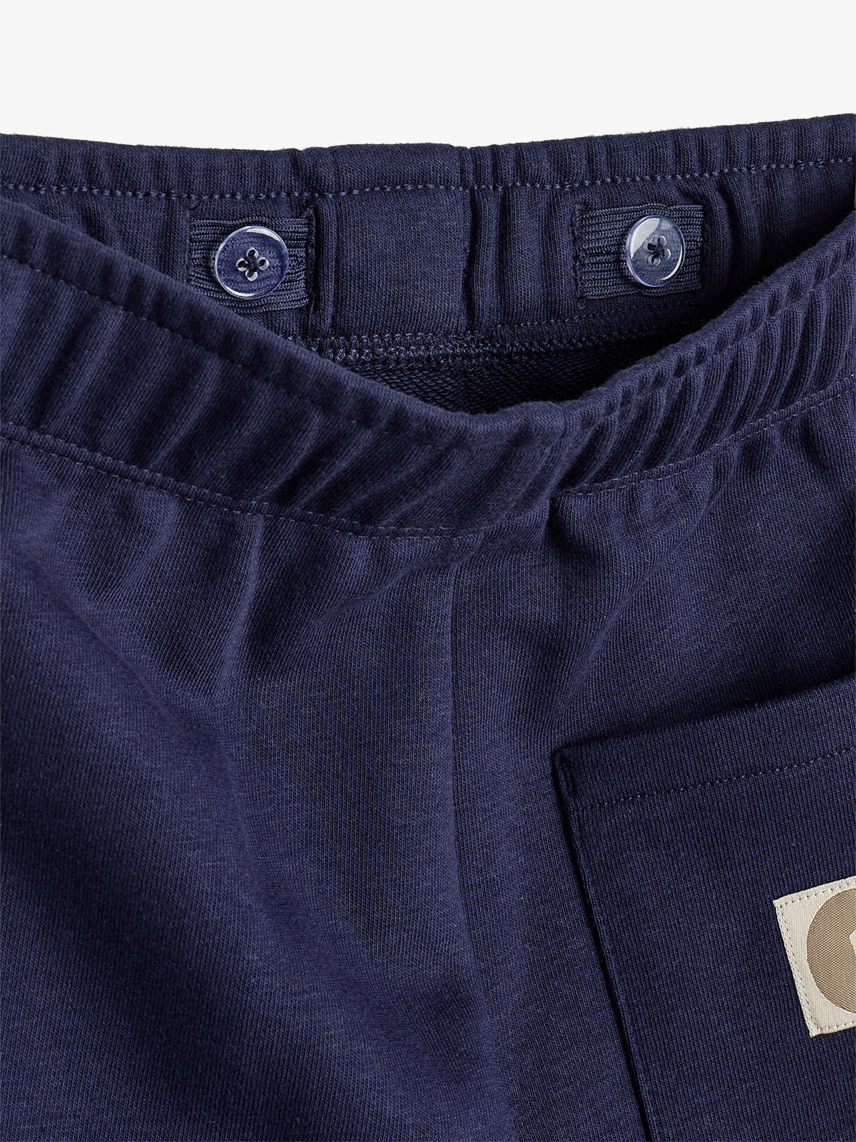 Navy blue pants with an elastic waistband and button adjustments. A pocket with a beige patch is visible, displaying a partial letter "G."