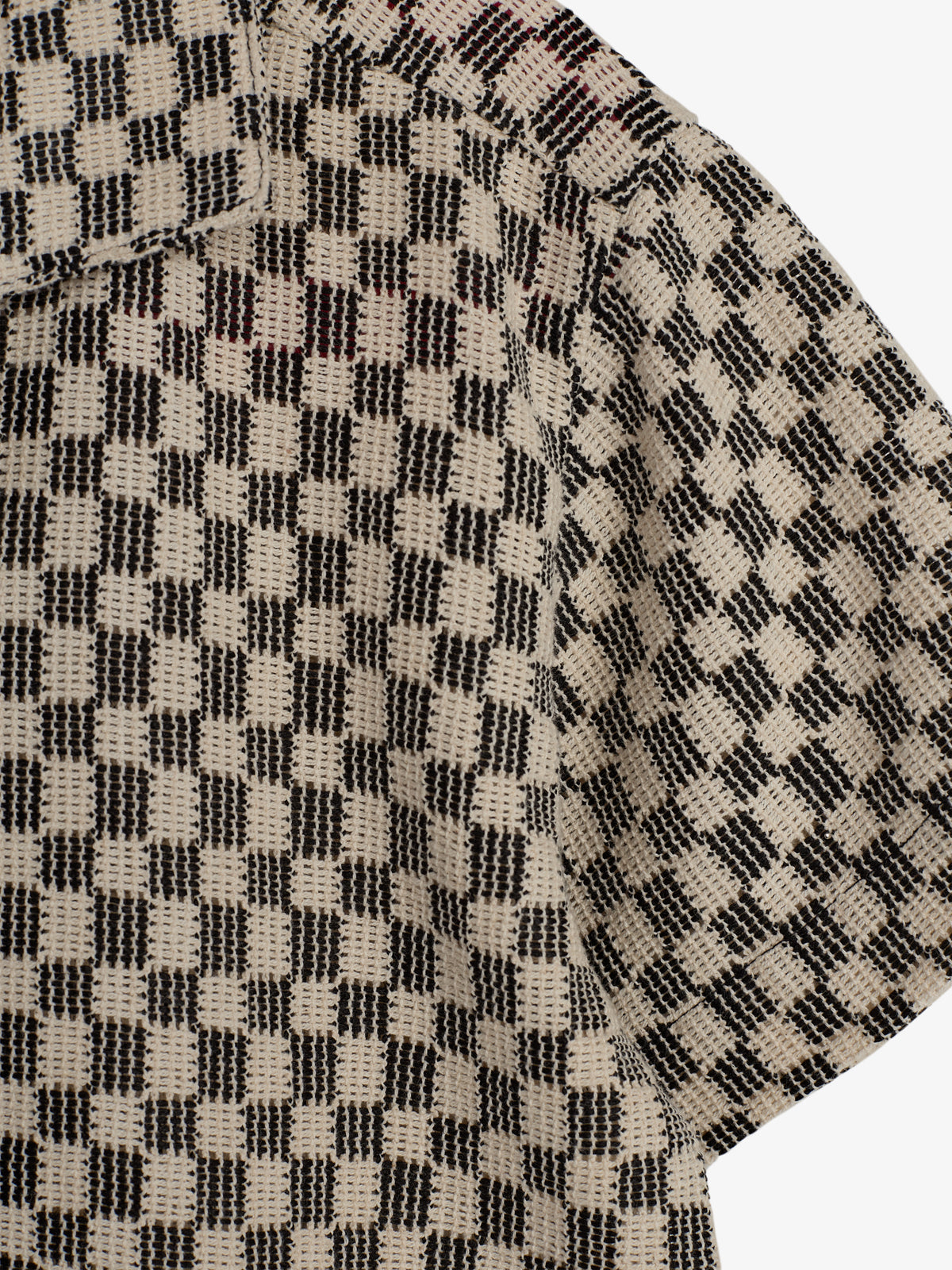 A close-up of a textured fabric with a black and beige checkered pattern. It features a visible seam and part of a short sleeve, suggesting it's part of a garment.