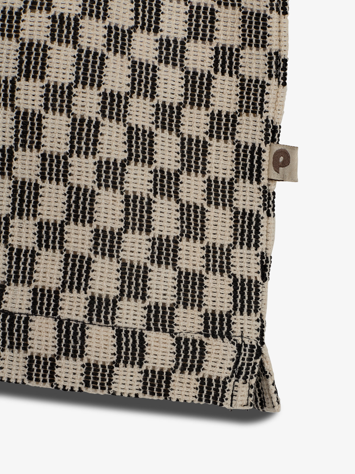 A checkered fabric in black and beige with a textured pattern, featuring a small tag with the letter "P" on the side, against a plain white background.