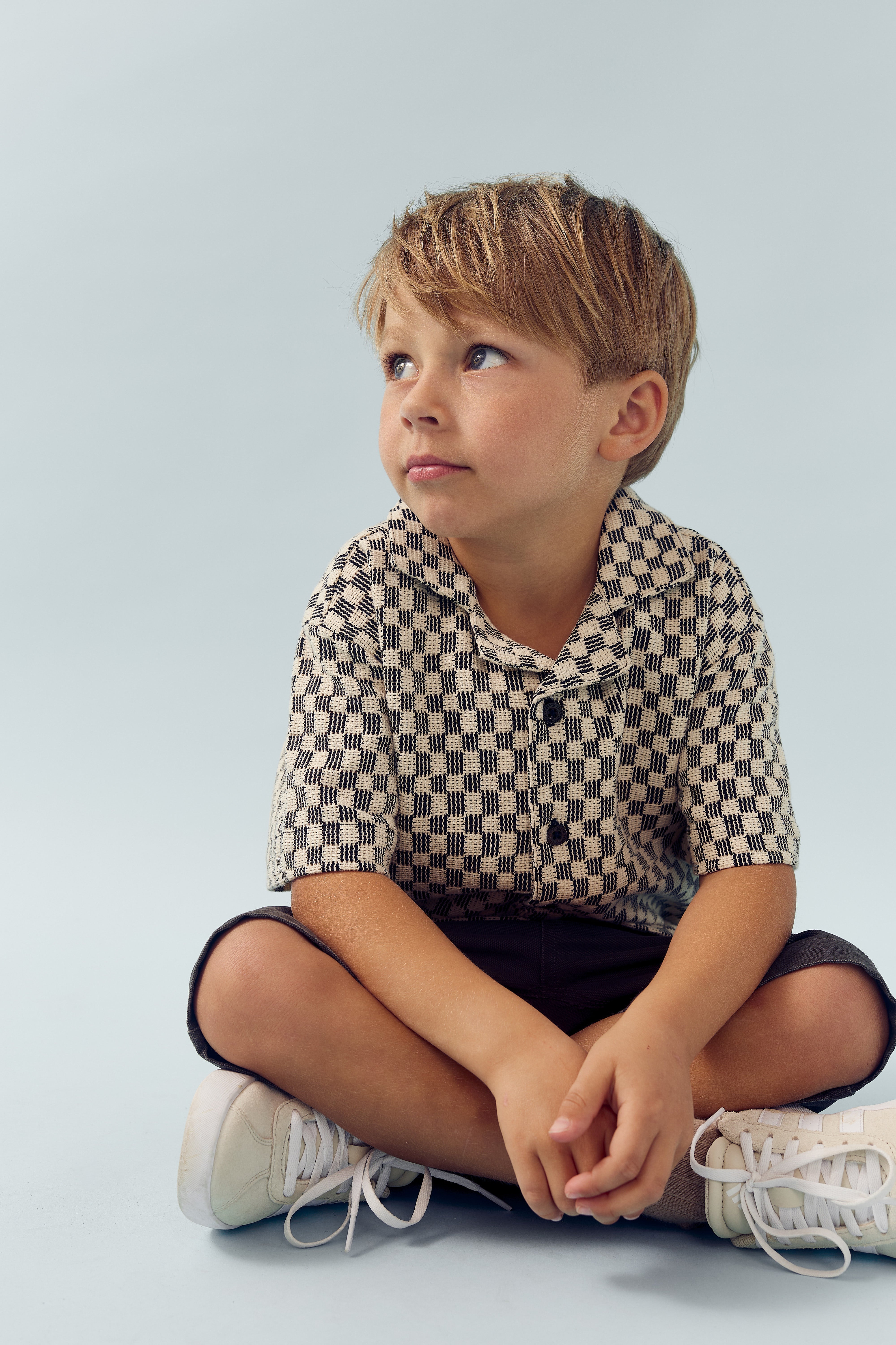 A child sits cross-legged, wearing a checkered shirt, dark shorts, and sneakers, gazing thoughtfully to the side against a plain, light blue background.