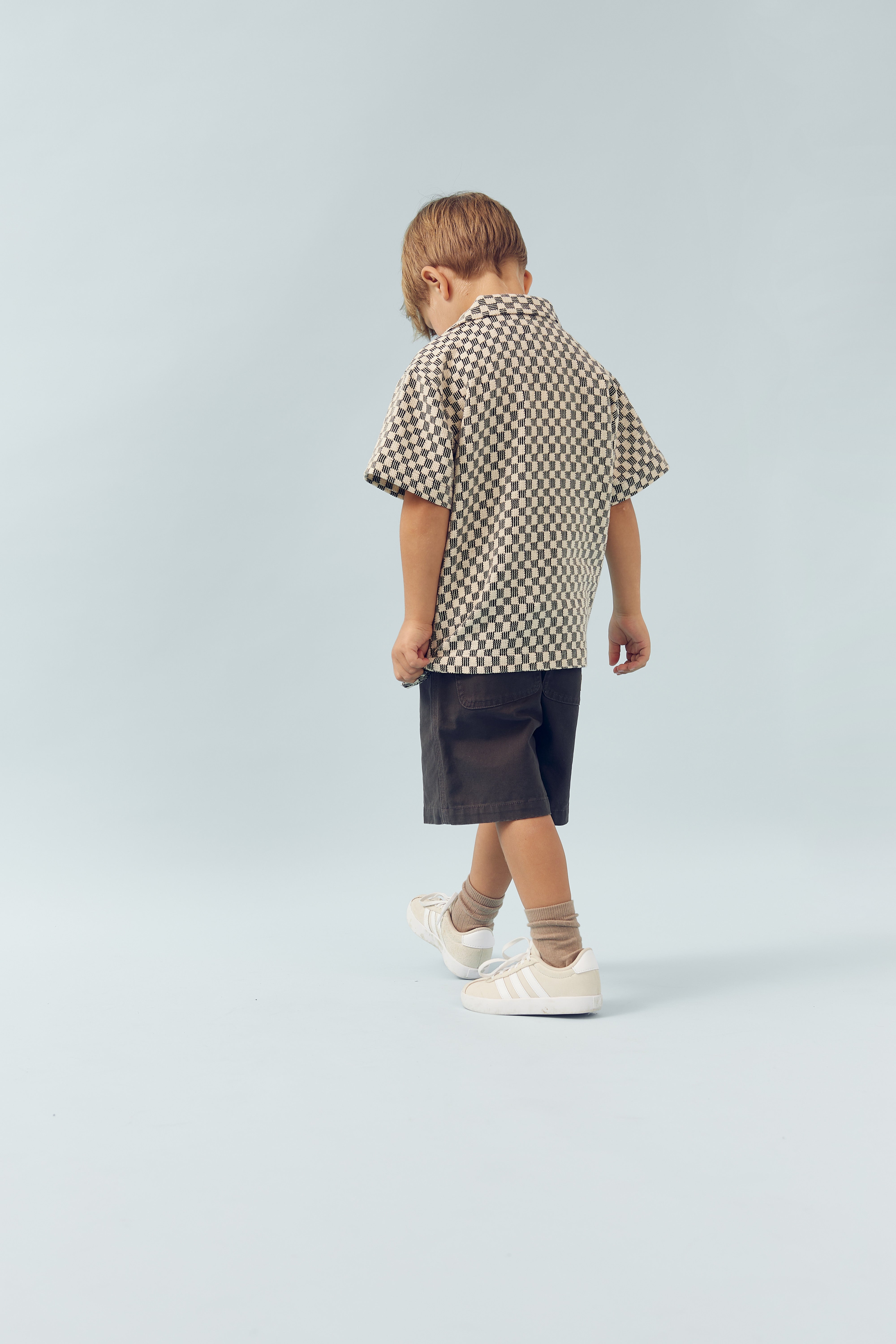 A child dressed in a checkered shirt and dark shorts walks in a spacious, light-colored room, wearing white sneakers and beige socks.