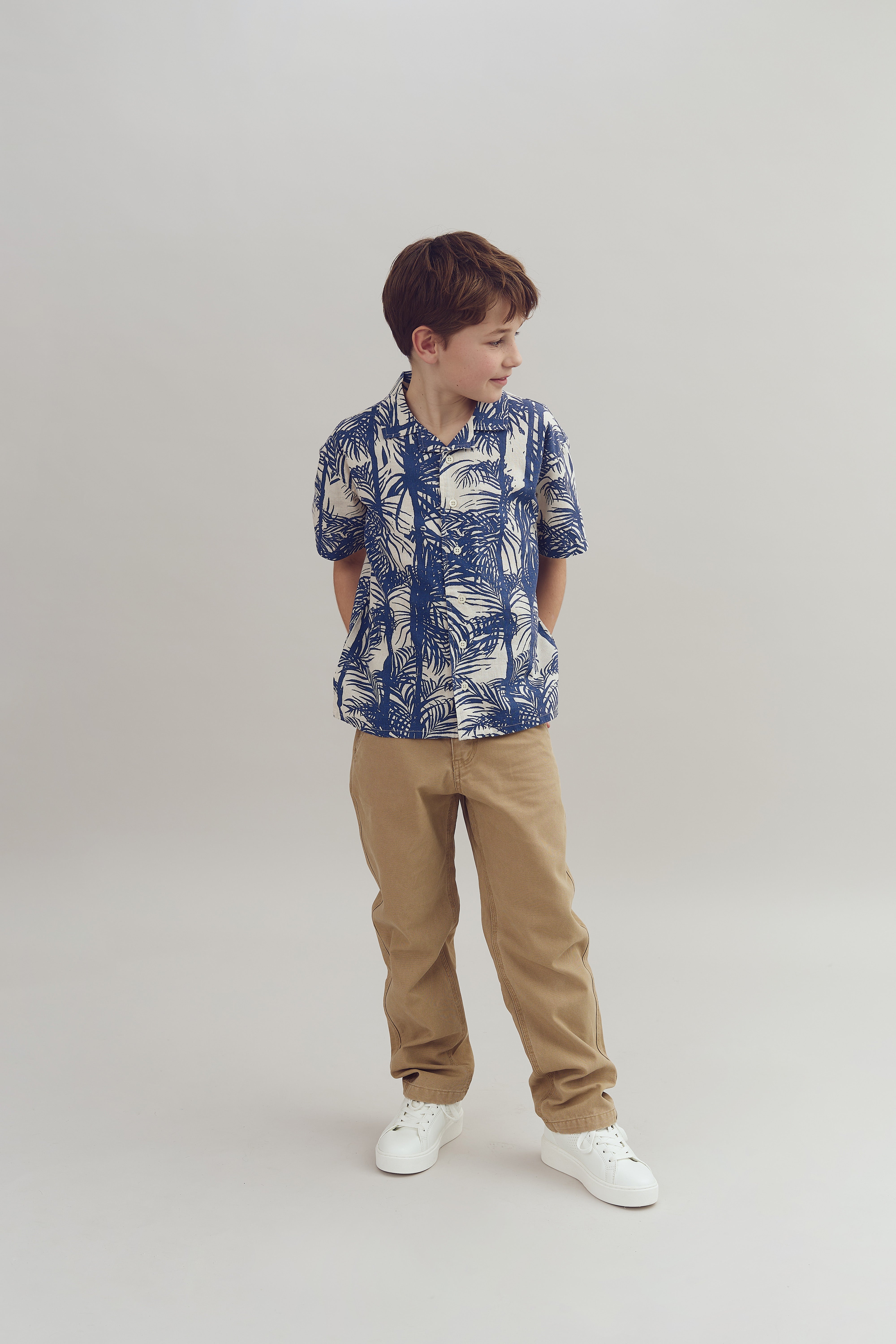 A child stands with hands behind back, wearing a blue and white patterned shirt and beige pants, looking sideways in a studio setting with a plain background.