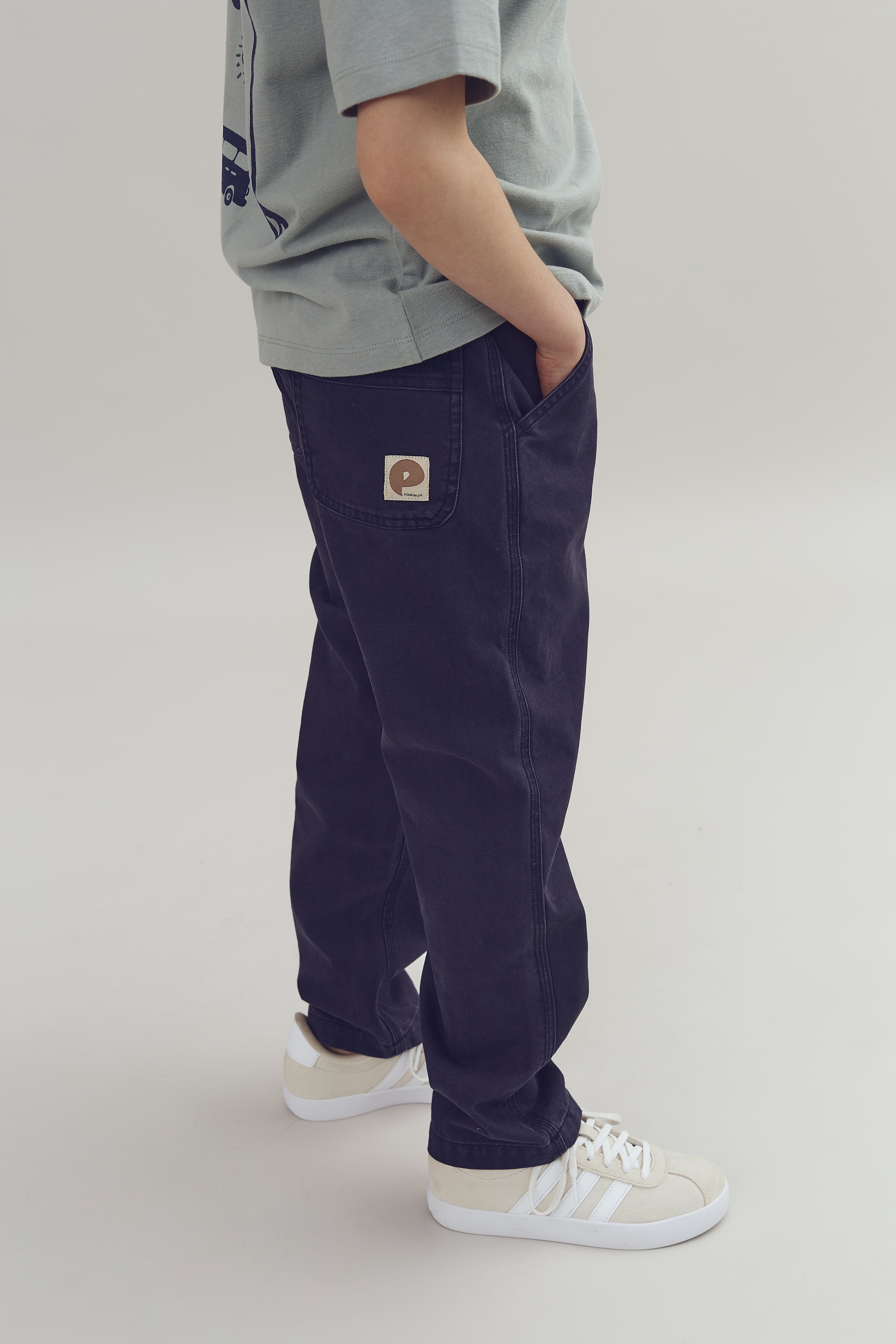 A child in a light gray shirt and navy pants with one hand in the pocket, stands on a plain background wearing white sneakers. A patch with "PSA FIRE" is visible.
