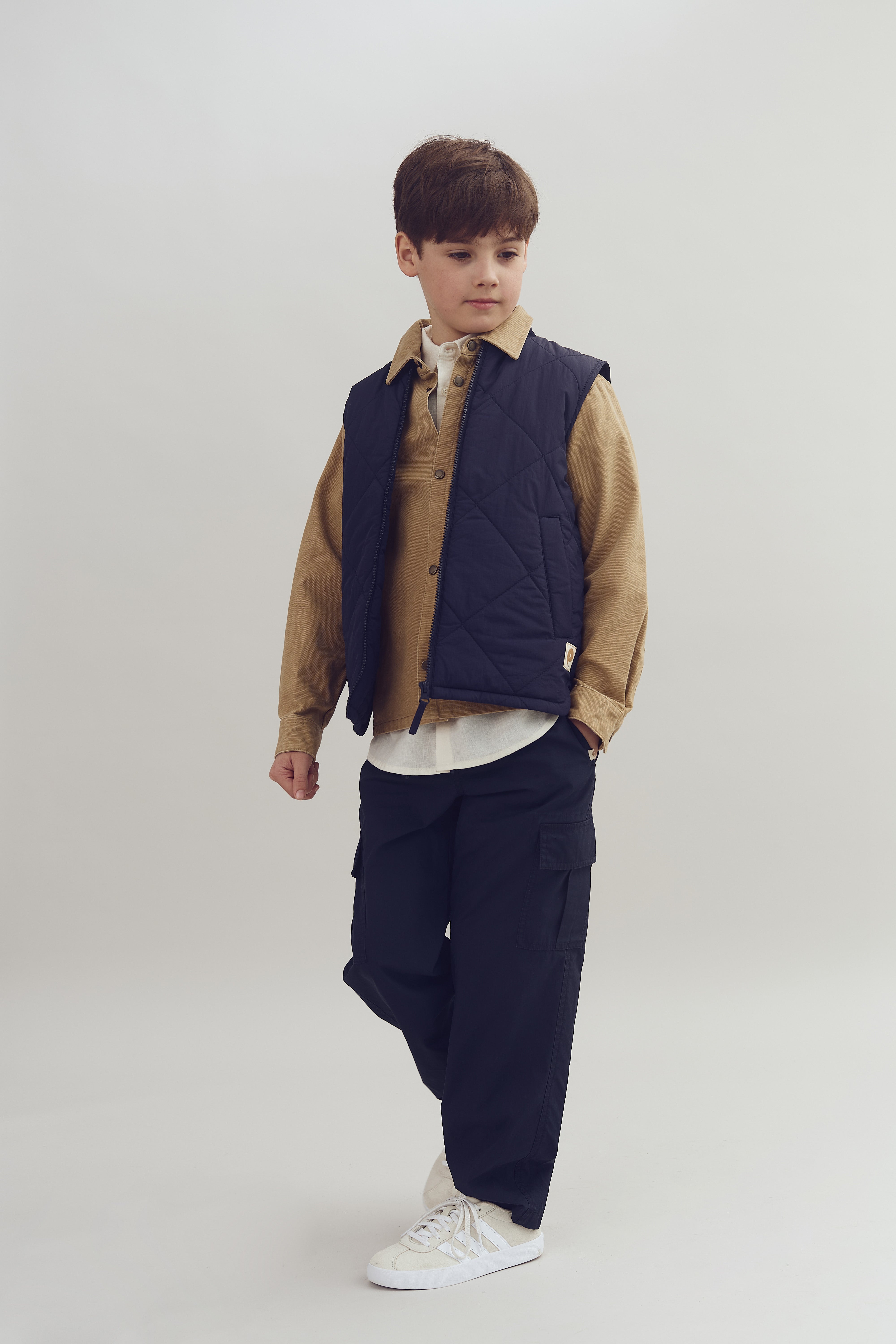 A child stands wearing a navy quilted vest over a tan shirt, dark cargo pants, and white sneakers. The background is a neutral, light color.