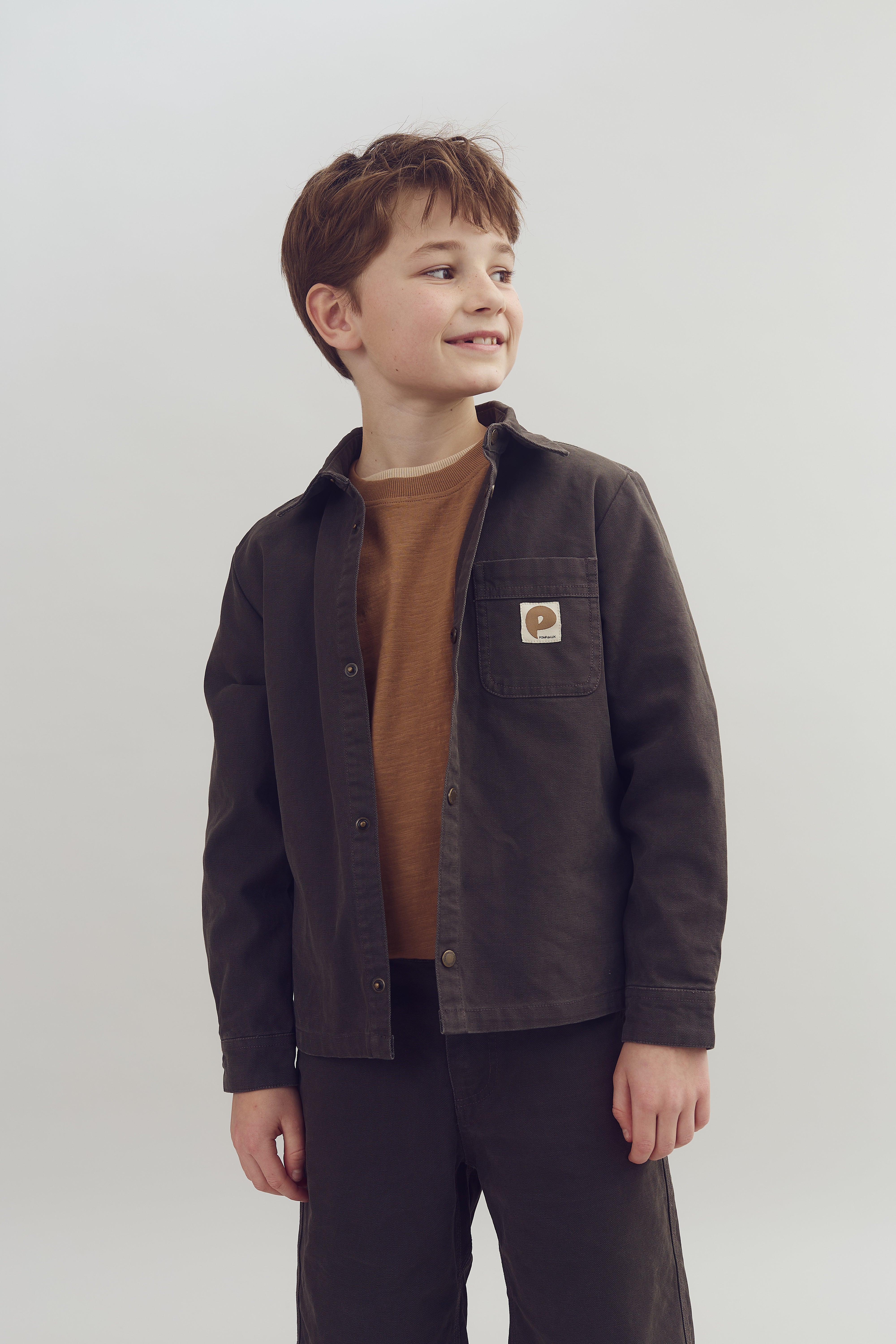 A child stands wearing a brown jacket with a "p-opar" patch on the pocket over a knitted sweater, looking to the side against a plain background.