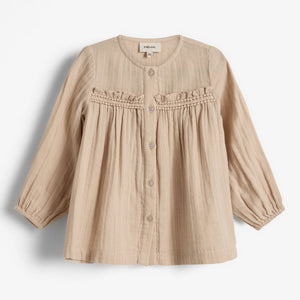 Beige kinderoverhemd met knopen en geplooide details aan de bovenkant, hangend aan een witte achtergrond. Etiket "pure.love." met maat "18M" zichtbaar binnenin.