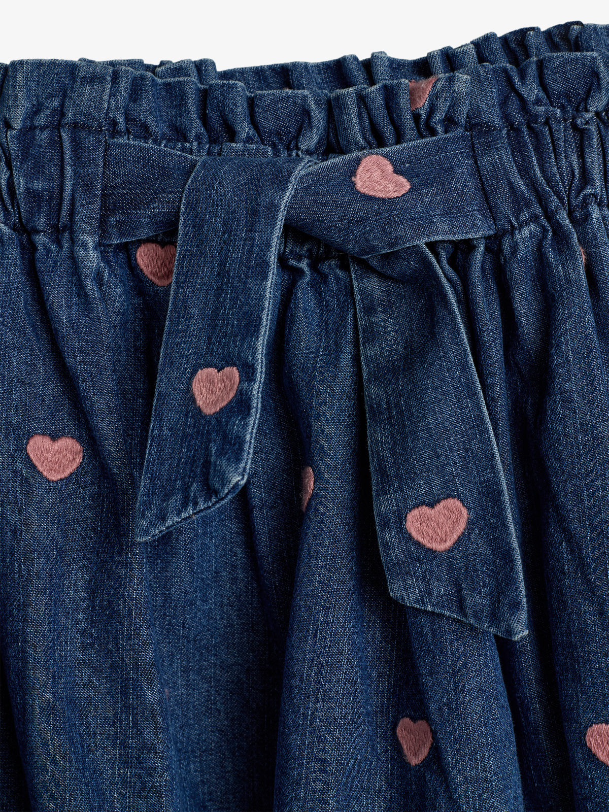 A denim skirt features pink heart patterns and a tie at the waistband, with gathering along the top edge, creating a textured appearance against the plain background.