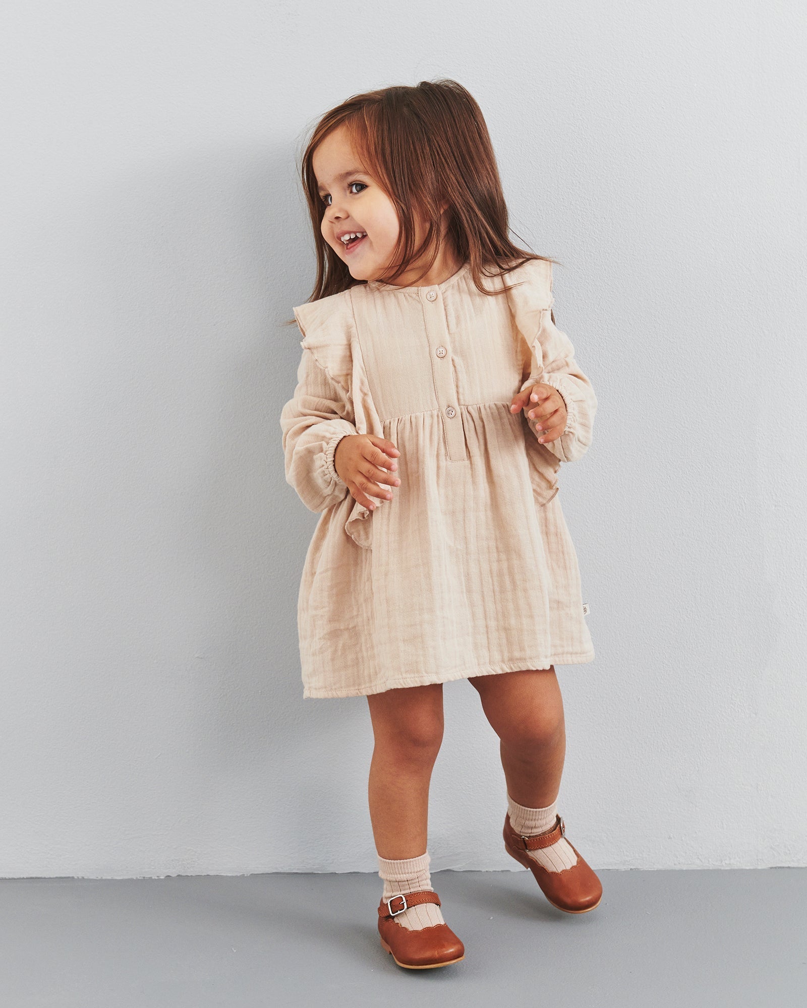 Een jong meisje draagt een beige jurk en bruine schoenen, lachend terwijl ze tegen een lichtgrijze muur staat. Haar lange haar hangt los over haar schouders.