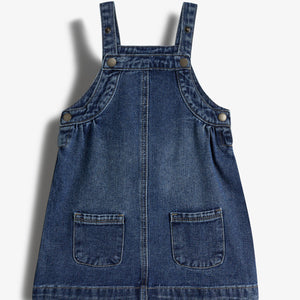 Denim pinafore-jurk, onbeweeglijk op afbeelding, met twee zakken en schouderbandjes met knopen. Geen tekst zichtbaar. De context is een effen witte achtergrond.