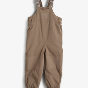 Een kinderregenbroek met verstelbare schouderbanden en elastische zomen. De broek is bruin en heeft een cirkelvormig logo met de tekst "OMP DELUX ADVENTURE" op de borst.