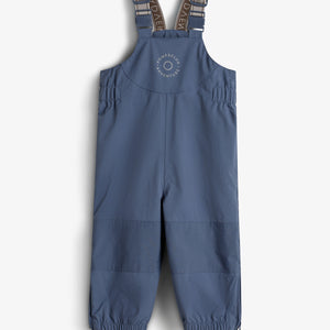 Een blauwe kinderregenbroek met verstelbare schouderbanden en elastische enkels, ontworpen voor avonturen. Op de borst staat de tekst “ROMPDELUX ADVENTURE”. Het hangt tegen een neutrale achtergrond.