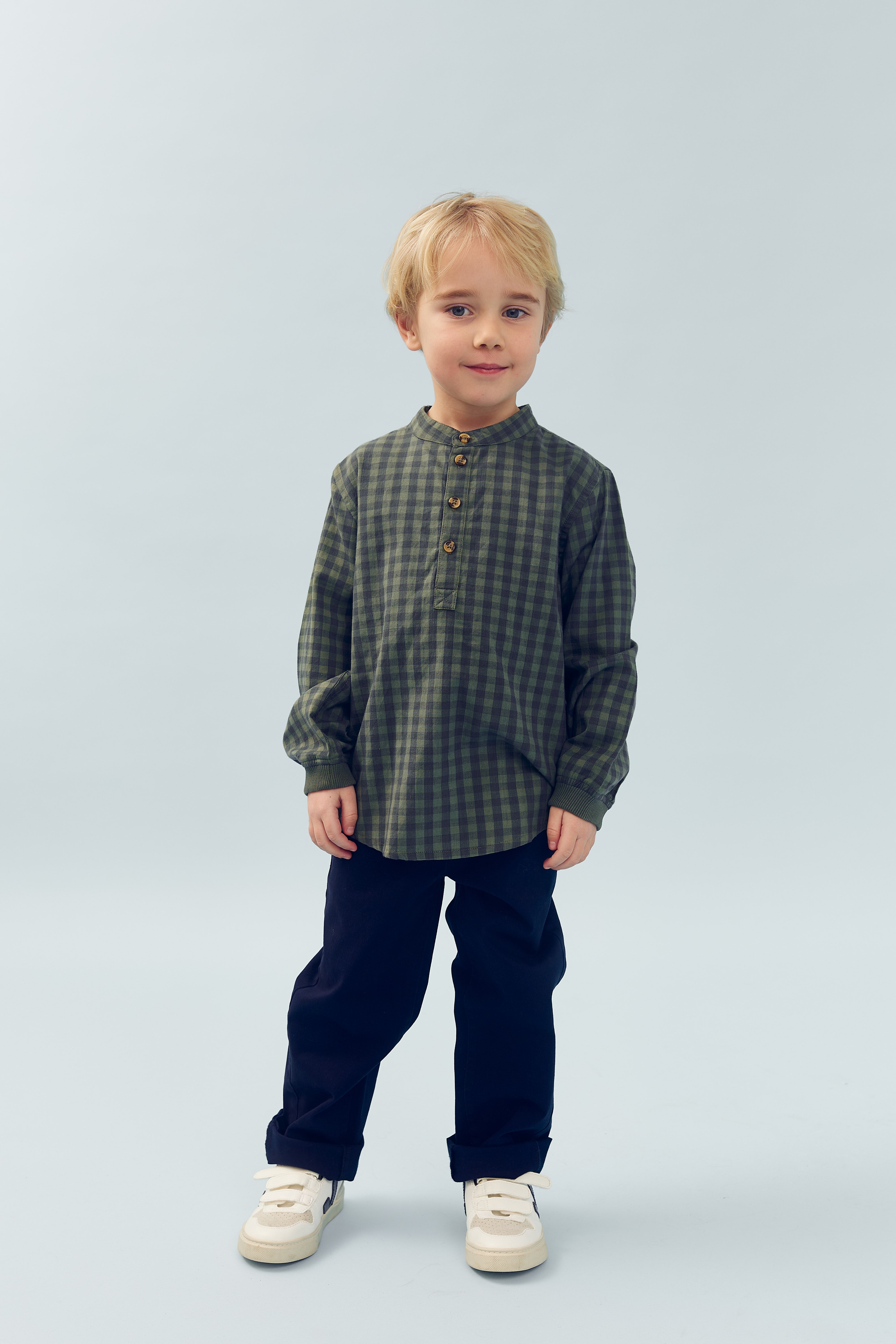 Een kind draagt een ruitjesoverhemd en zwarte broek, staat rechtop en kijkt vriendelijk, in een neutrale, lichtgekleurde achtergrond. Er is geen tekst zichtbaar.
