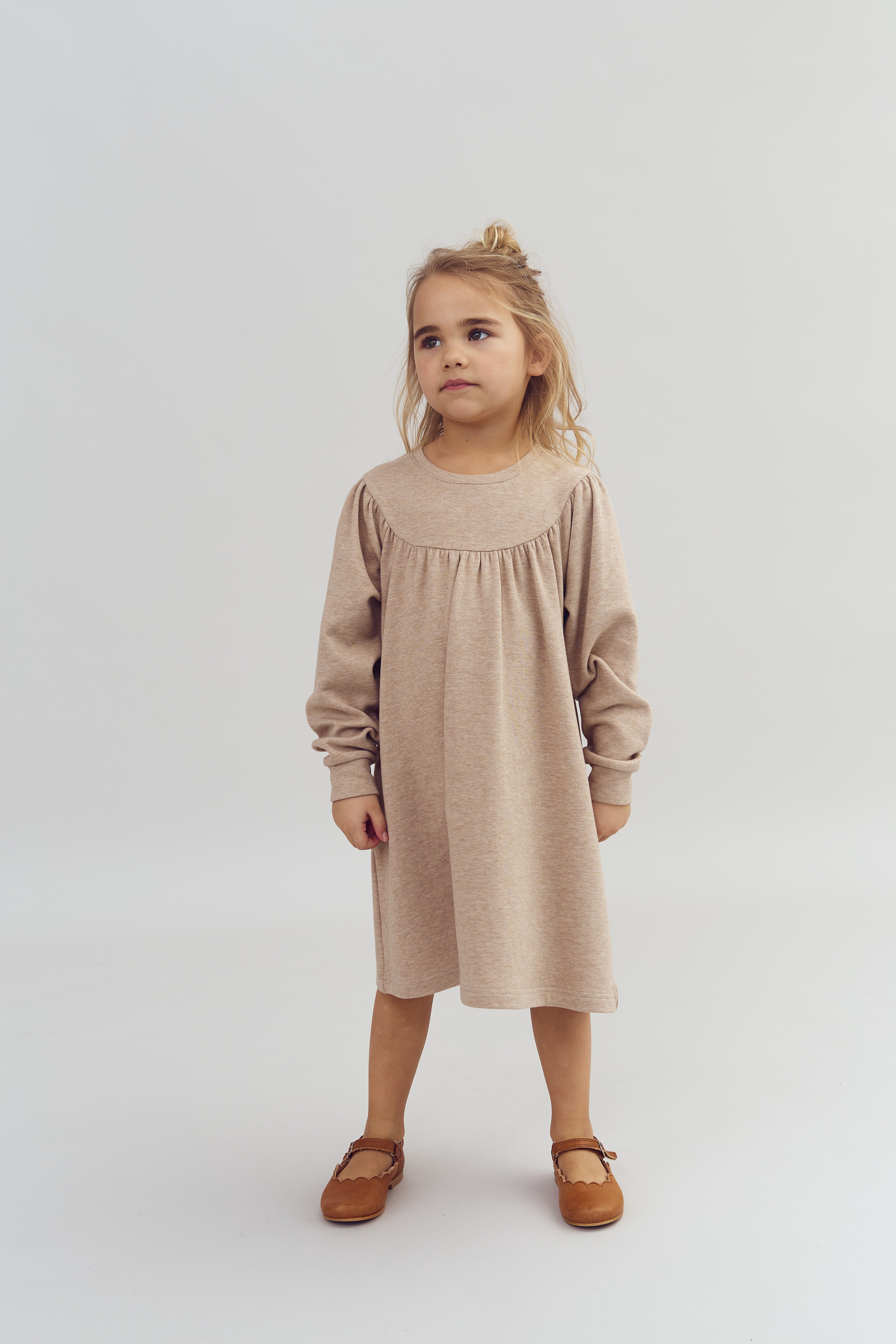 Kind in beige jurk staat rechtop, kijkt rustig voor zich uit. De achtergrond is egaal wit, waardoor de aandacht op het kind gericht is.