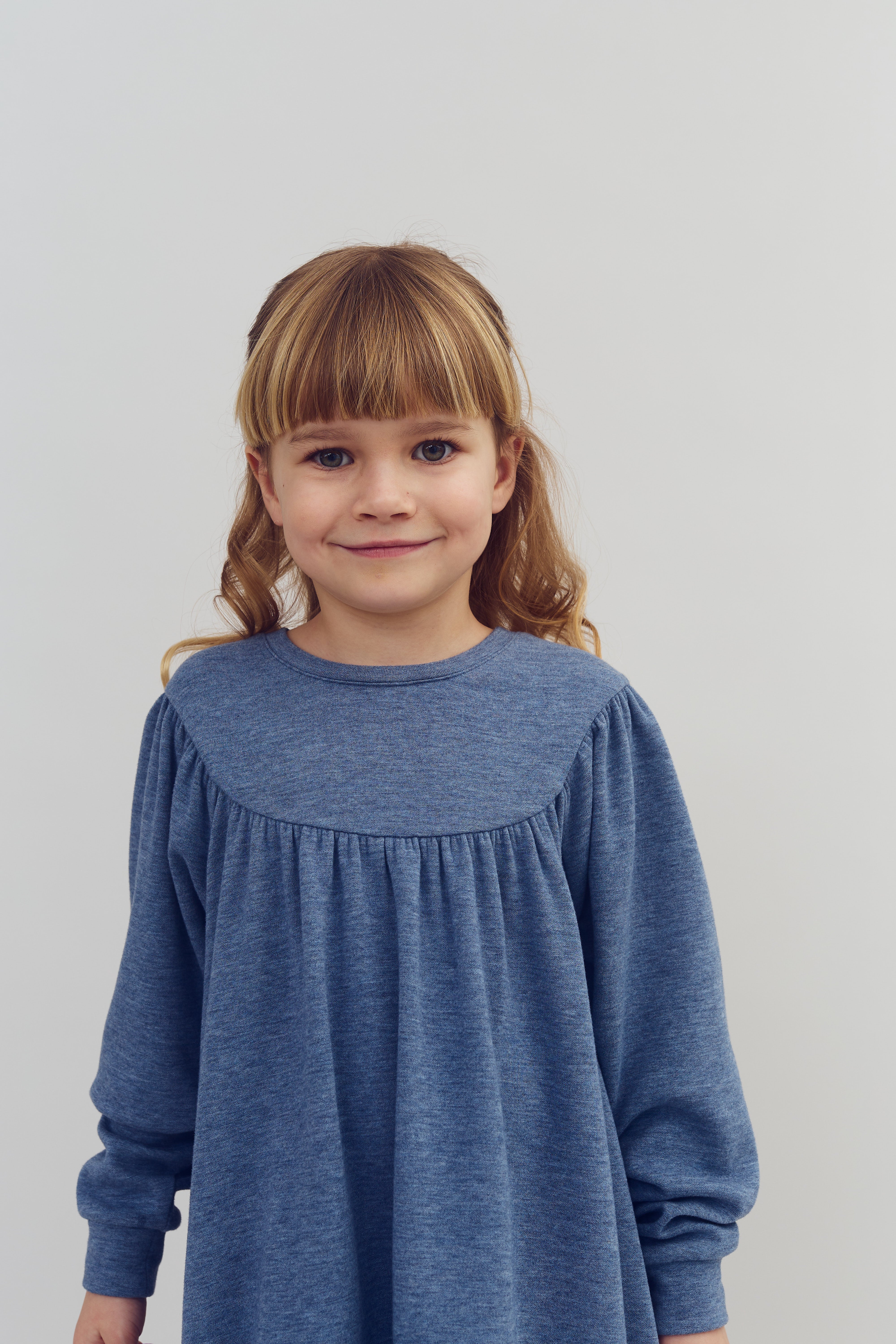 Een meisje in een blauwe jurk glimlacht in een neutrale omgeving met een effen lichtgrijze achtergrond. Haar blonde haar is gestyled met krullen aan de uiteinden.