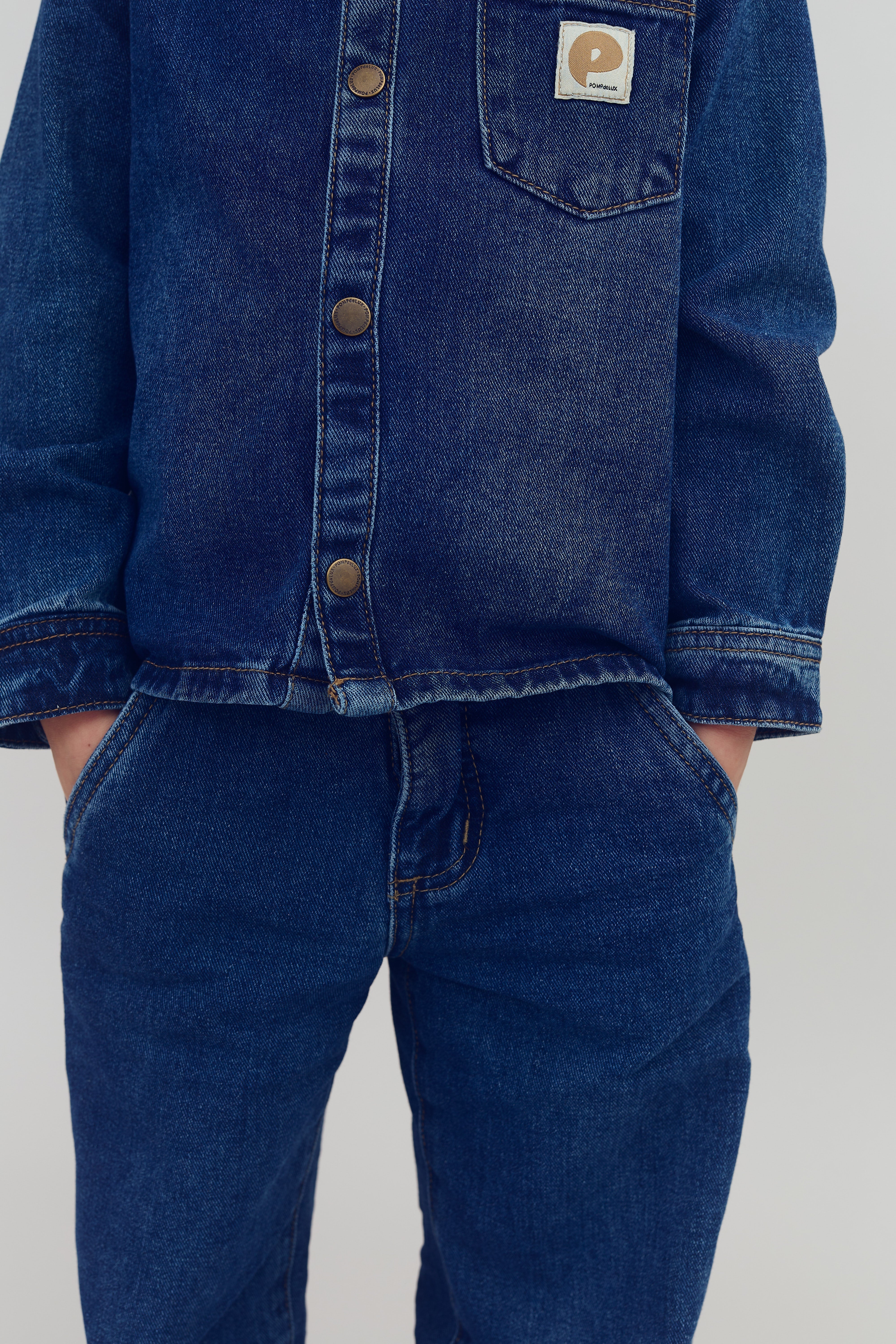Een persoon draagt een blauwe spijkerjas en -broek, handen in de zakken. Op de jas zit een label met de tekst "P". De achtergrond is effen lichtgrijs.