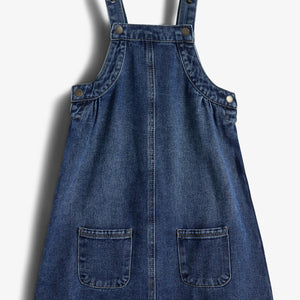 Blauwe denim jurk met schouderbanden en zakken aan de voorkant, liggend op een witte achtergrond. Bevestigingsknopen zijn zichtbaar op de bandjes en zijkanten.