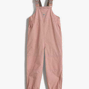 Een roze kinderregenbroek met verstelbare schouderbanden op een witte achtergrond. De tekst op de bandjes is niet volledig leesbaar. Elastische enkelmanchetten en voetbanden zijn zichtbaar.