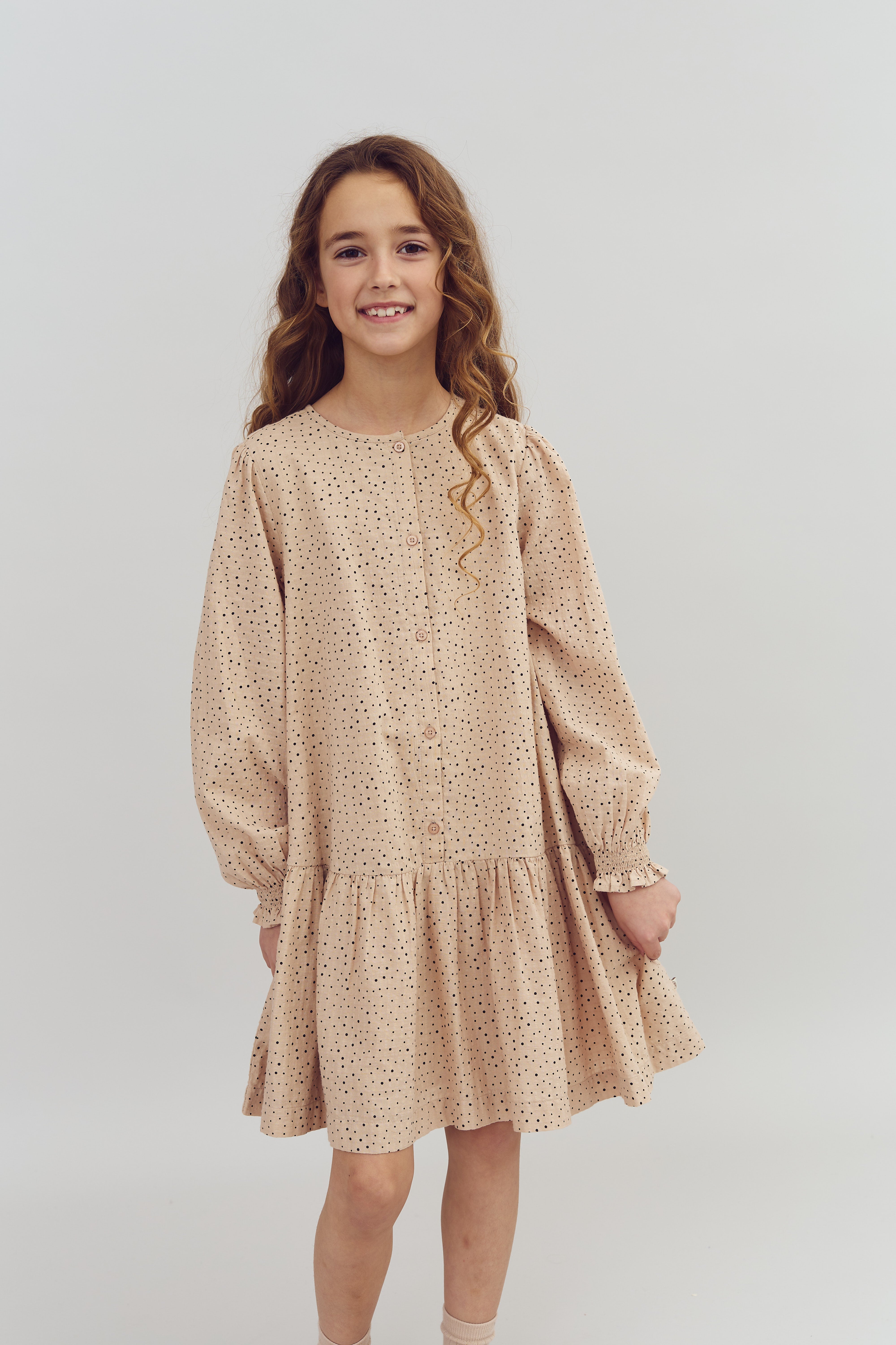 Meisje glimlacht, draagt een beige jurk met zwarte stippen, lange mouwen en knopen. Ze heeft krullend haar. Achtergrond is een effen, lichte muur.