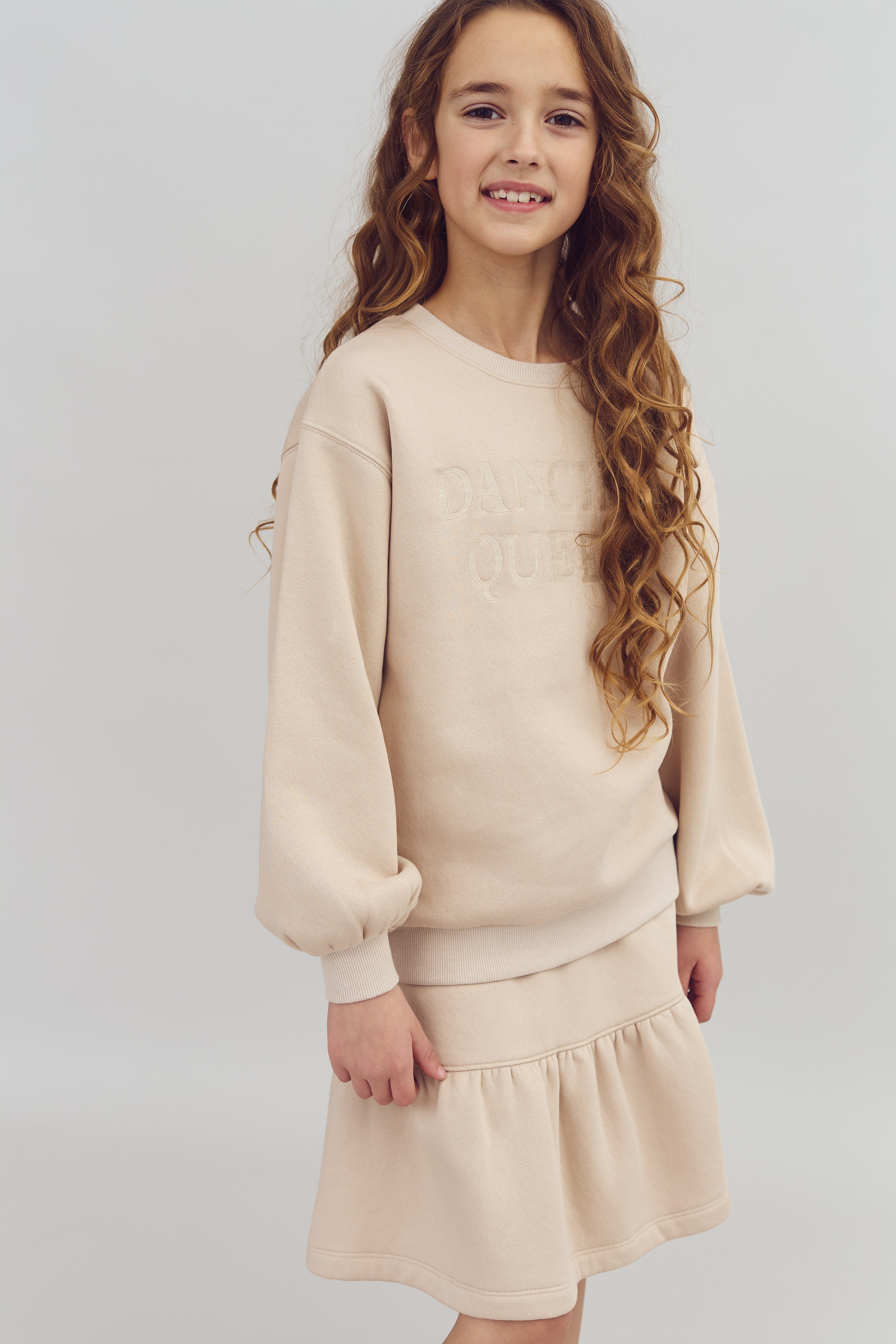 Een meisje poseert, draagt een beige trui met de tekst "DANC QUEE" in een eenvoudige studio-omgeving.