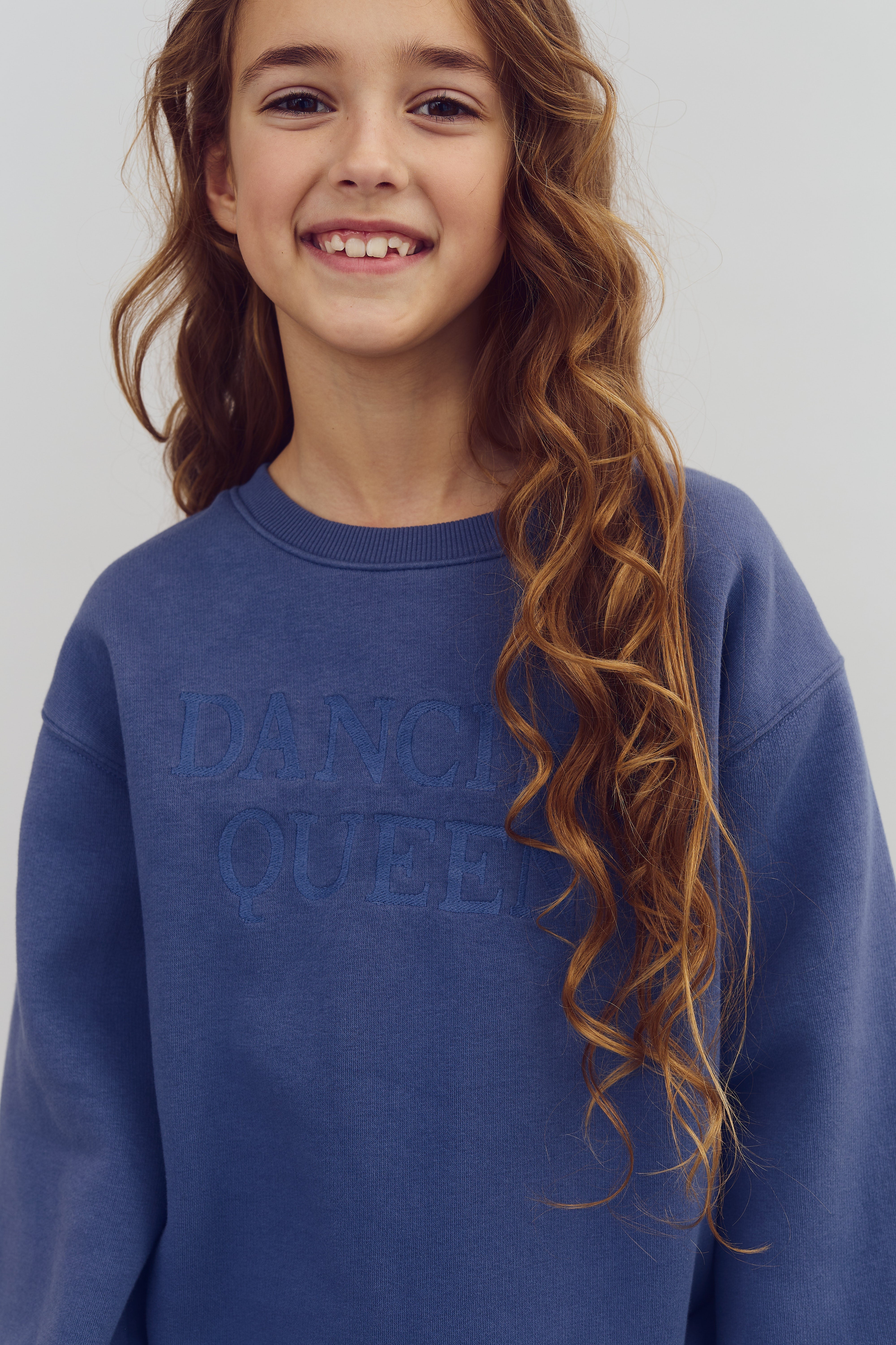 Een meisje glimlacht en draagt een blauwe trui met de tekst “DANCING QUEEN” in reliëf. Ze heeft lang, krullend haar en staat tegen een neutrale, lichte achtergrond.