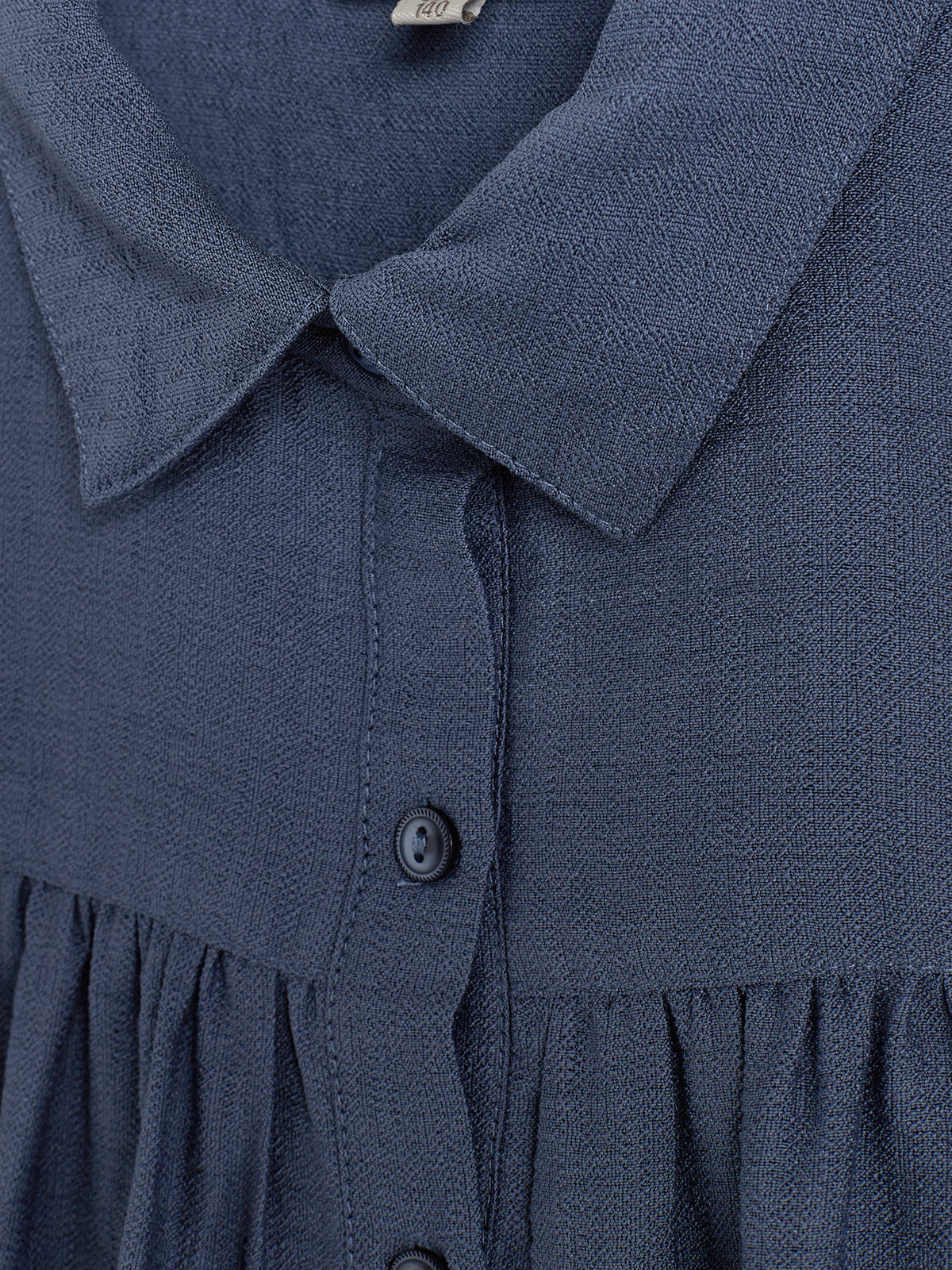 Donkerblauwe blouse met knoopsluiting en kraag, hangend tegen een neutrale achtergrond. Het materiaal heeft een textuur en een label met "140" is zichtbaar bij de kraag.