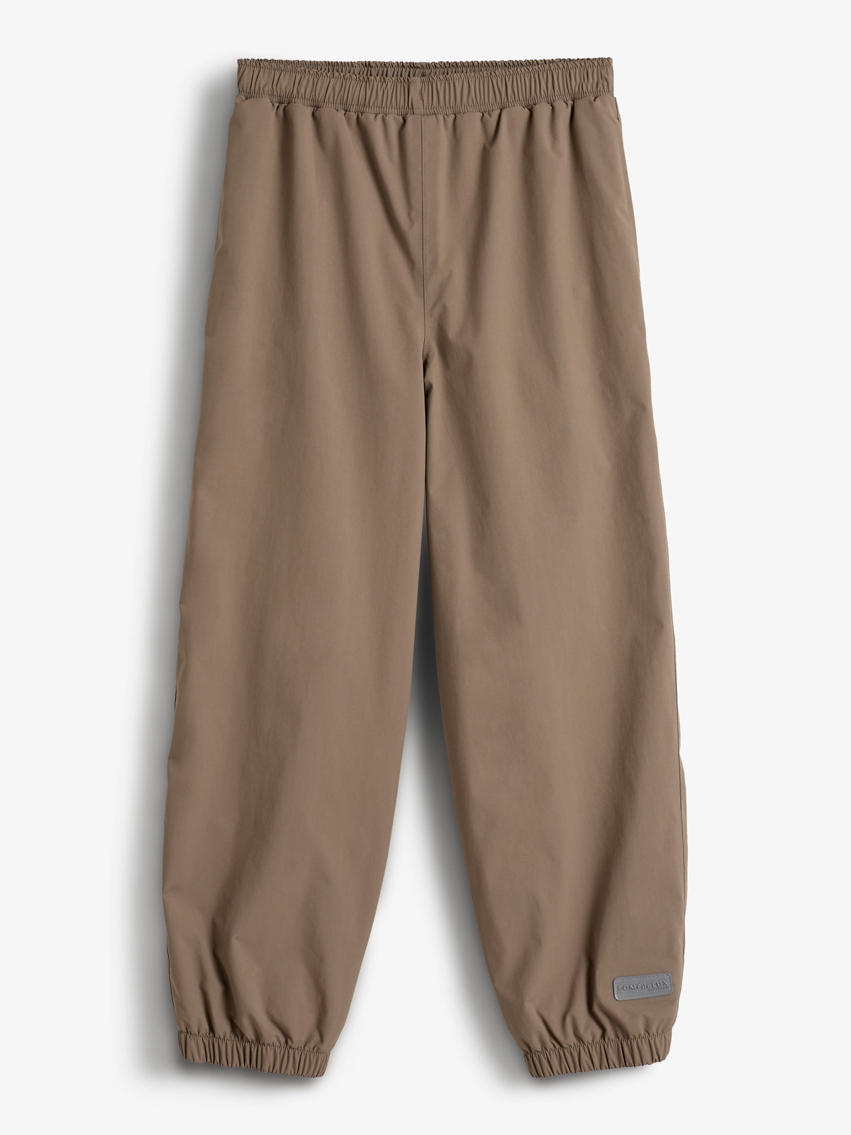 Een bruine broek hangt rechtop, met elastische tailleband en enkels. In een lichte, effen omgeving bevindt zich een label met de tekst "Polarn O. Pyret" rechtsonder.