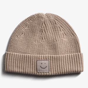 Een beige gebreide muts met een omgeslagen rand, waarop een label met een eenvoudig gezichtje en tekst "pOMPdeLUX" is bevestigd. De achtergrond is effen wit.