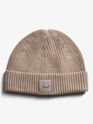 Een beige gebreide muts met een omgeslagen rand, waarop een label met een eenvoudig gezichtje en tekst "pOMPdeLUX" is bevestigd. De achtergrond is effen wit.