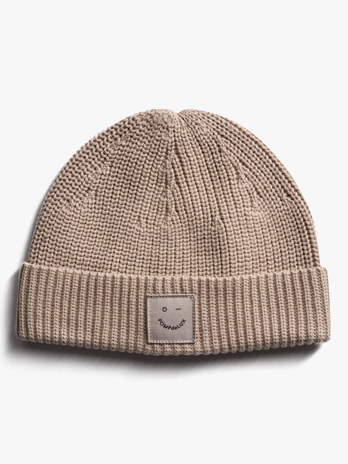 Een beige gebreide muts met een omgeslagen rand, waarop een label met een eenvoudig gezichtje en tekst "pOMPdeLUX" is bevestigd. De achtergrond is effen wit.