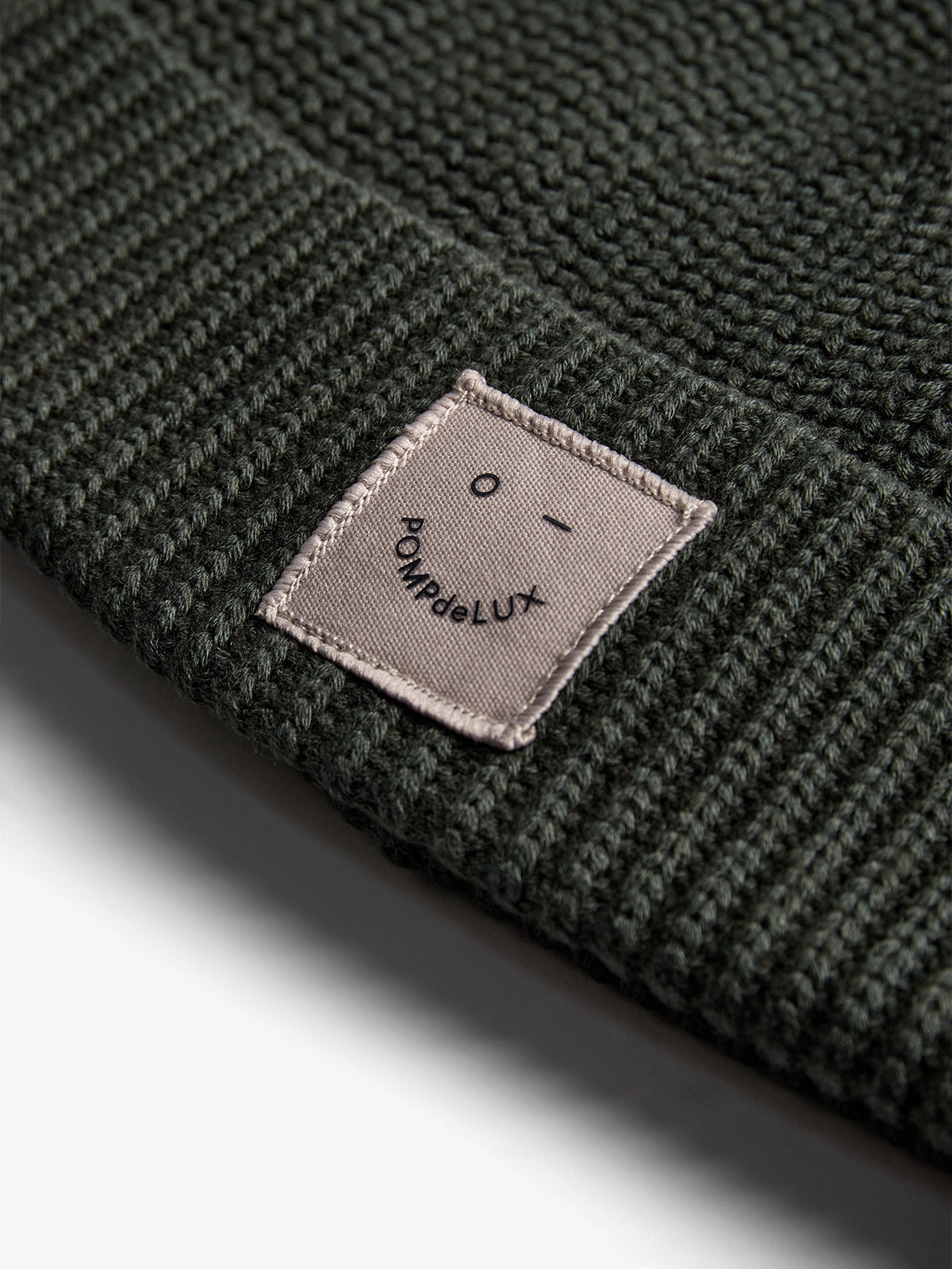 Een donkergroene gebreide muts met een beige rechthoekig label vastgenaaid aan de rand; tekst op het label: "POMPdeLUX" en een kleine smiley erboven.