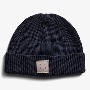 Een donkerblauwe gebreide muts ligt plat; er is een beige label met de tekst "POMPdeLUX" en een kleine glimlach. De achtergrond is effen wit.