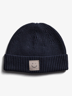 Een donkerblauwe gebreide muts ligt plat; er is een beige label met de tekst "POMPdeLUX" en een kleine glimlach. De achtergrond is effen wit.