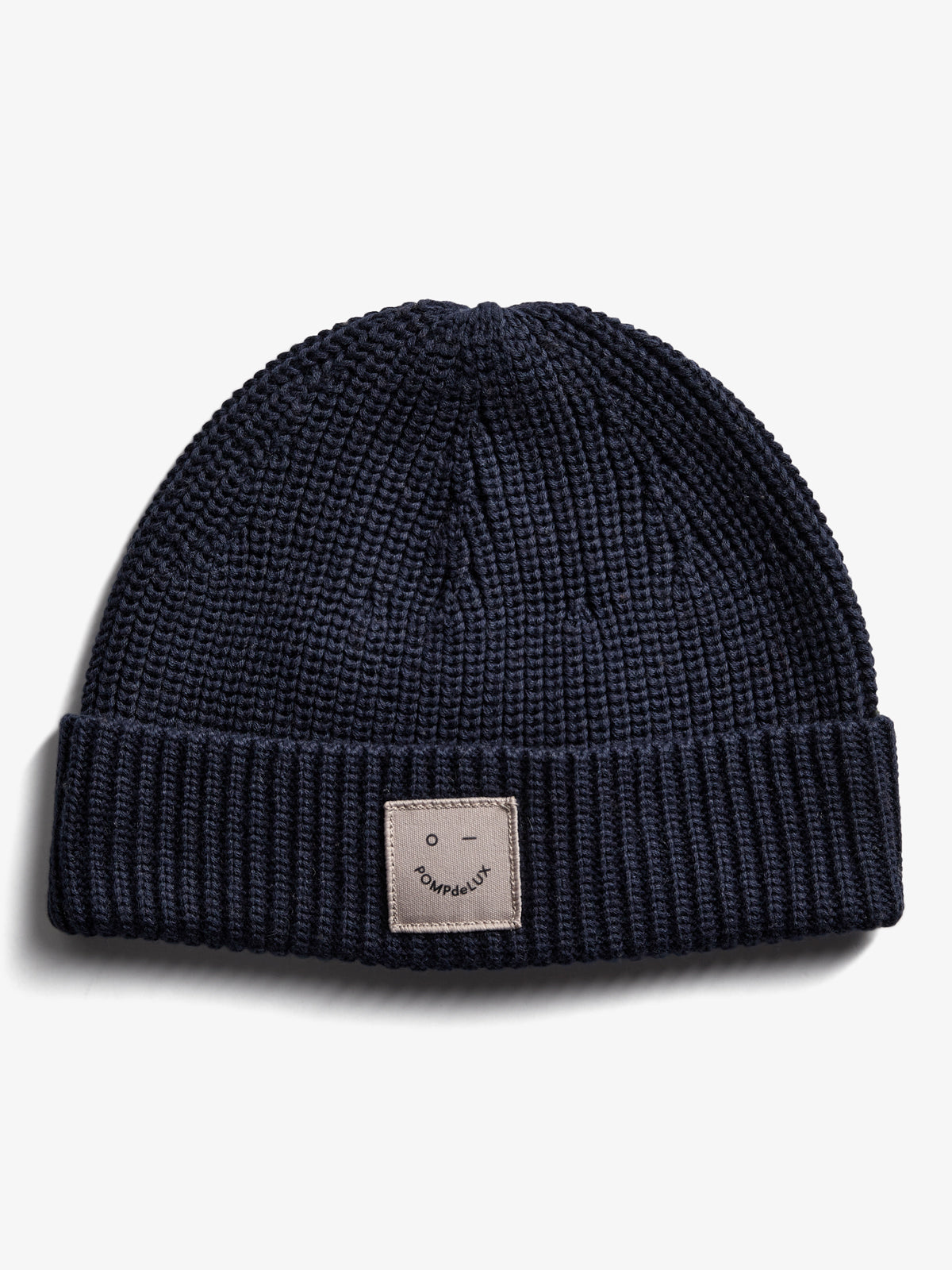 Een donkerblauwe gebreide muts ligt plat; er is een beige label met de tekst "POMPdeLUX" en een kleine glimlach. De achtergrond is effen wit.