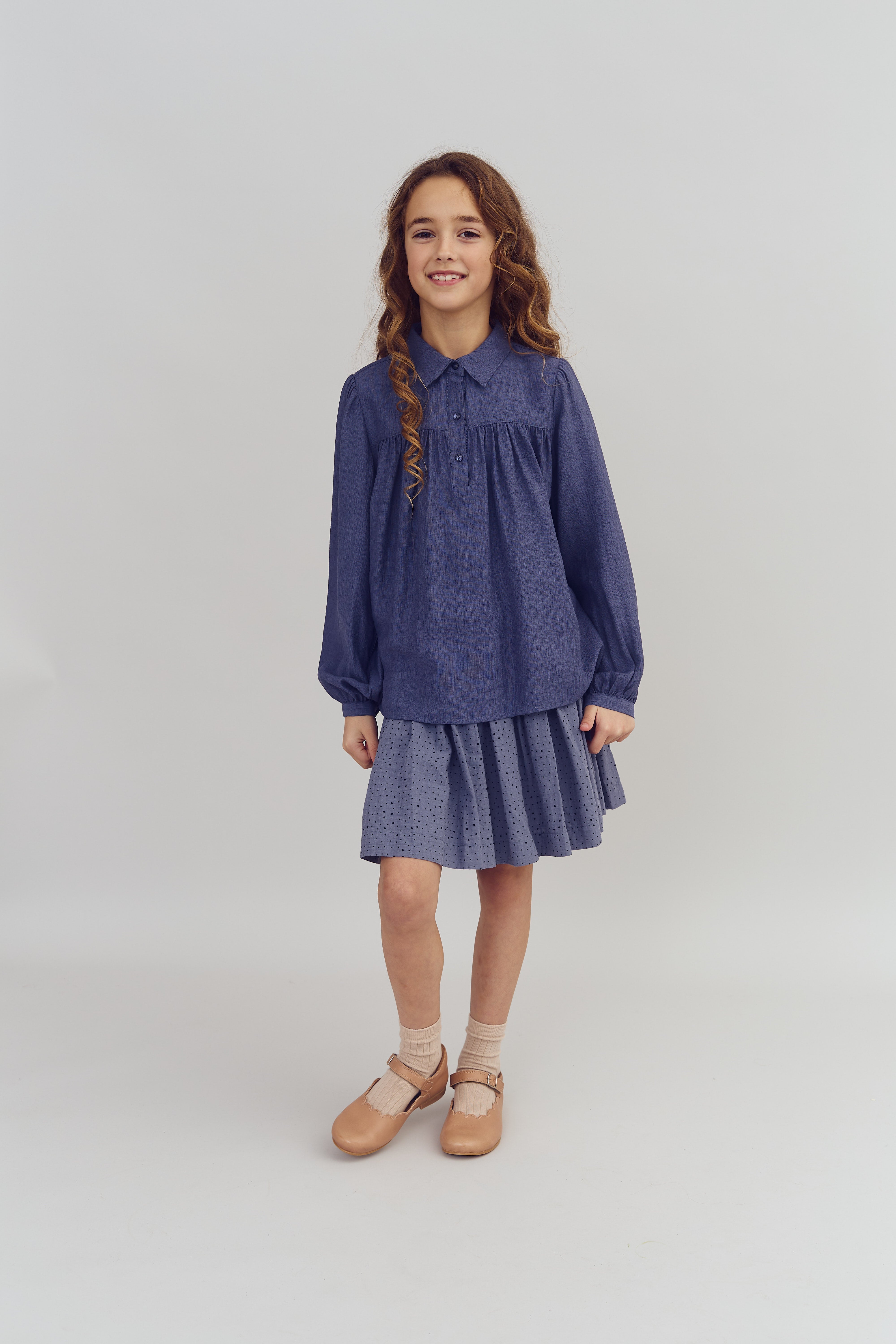 Een jong meisje staat glimlachend, gekleed in een blauwe blouse en rok, met bruine schoenen en sokken, op een neutrale, lichte achtergrond.