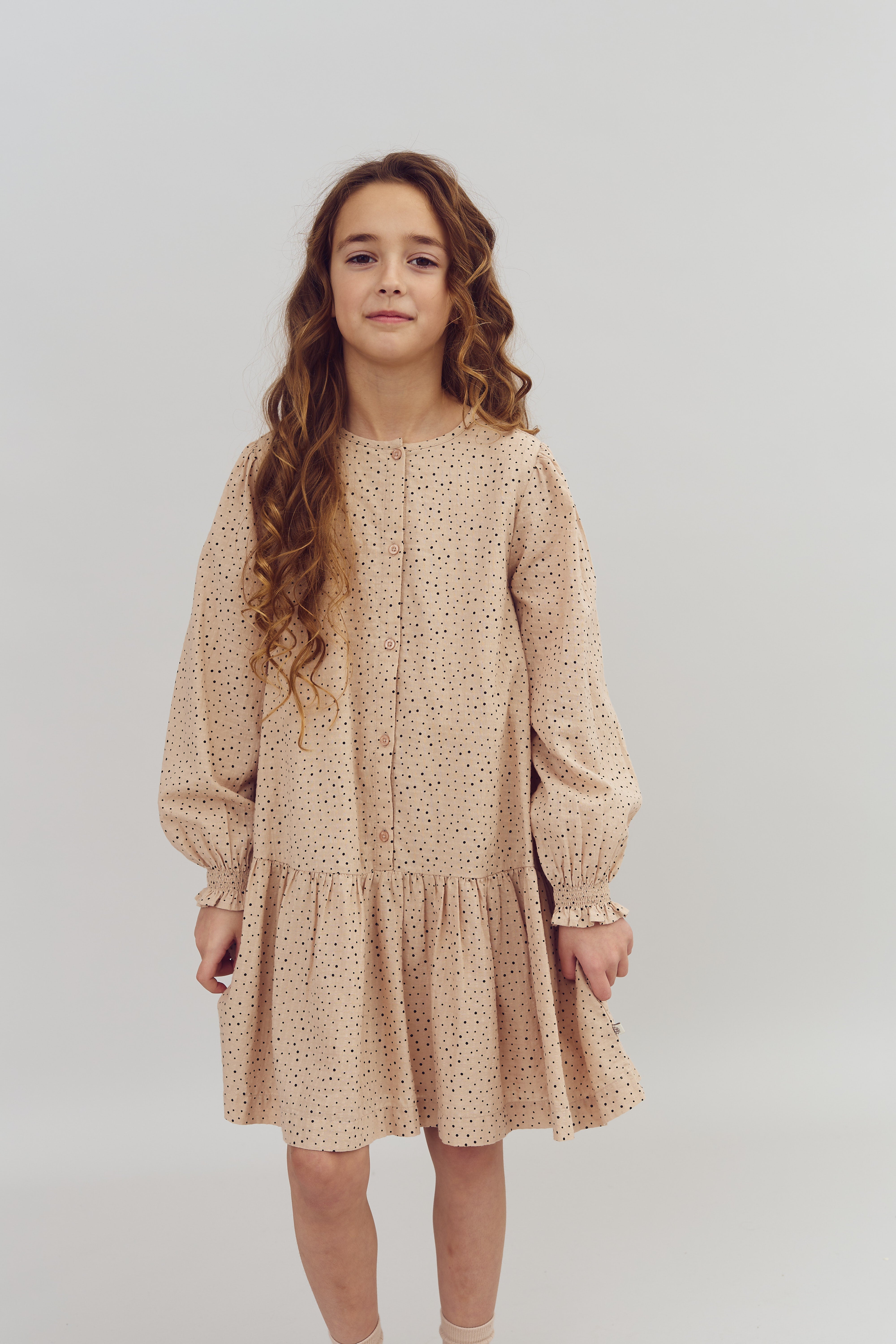 Meisje draagt een beige jurk met zwarte stippen, staat rechtop in een ruimte met een effen witte achtergrond. Ze heeft lang, krullend haar.