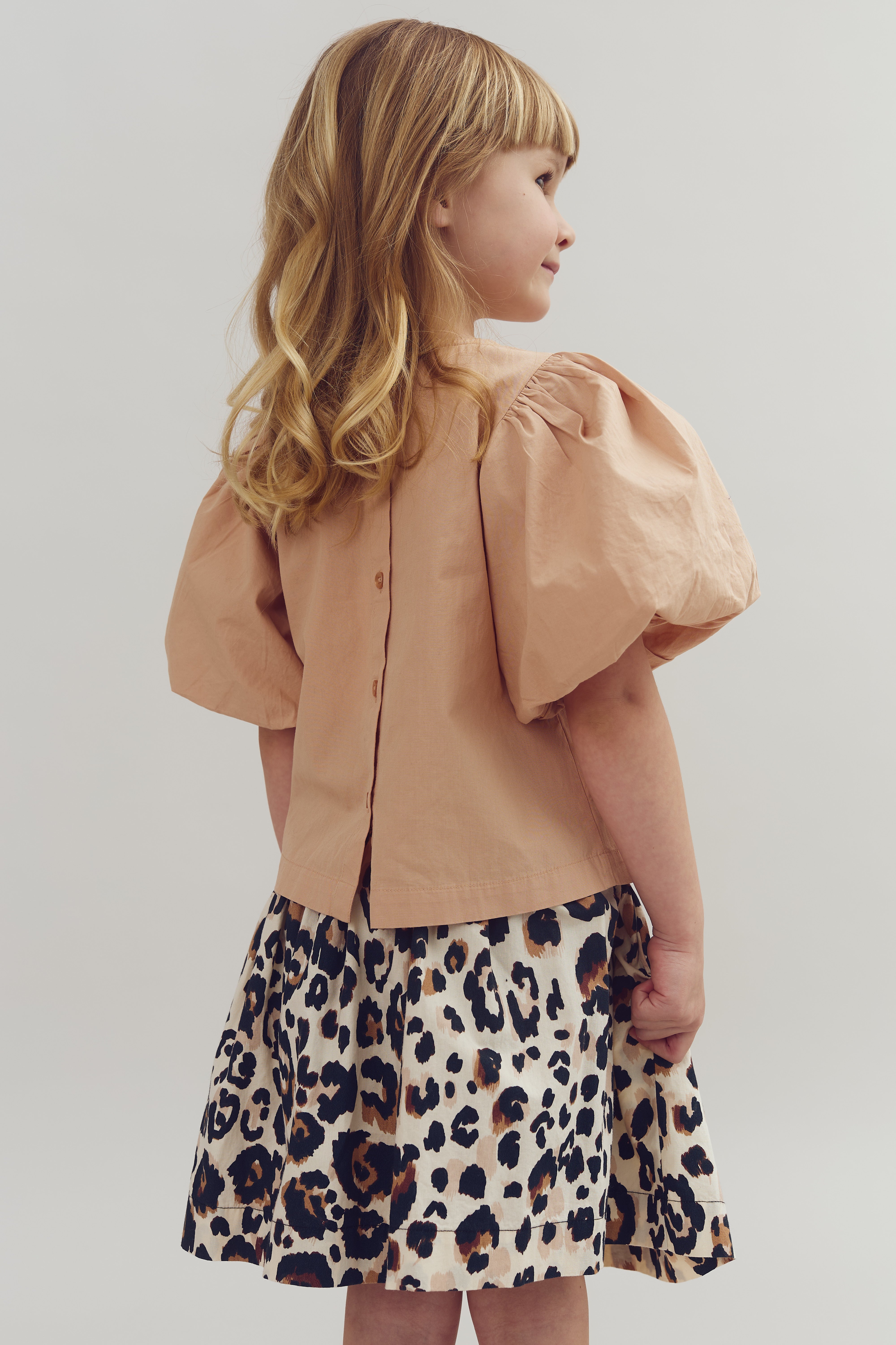 A child stands sideways wearing a peach puff-sleeve blouse and a leopard print skirt, against a plain light gray background.