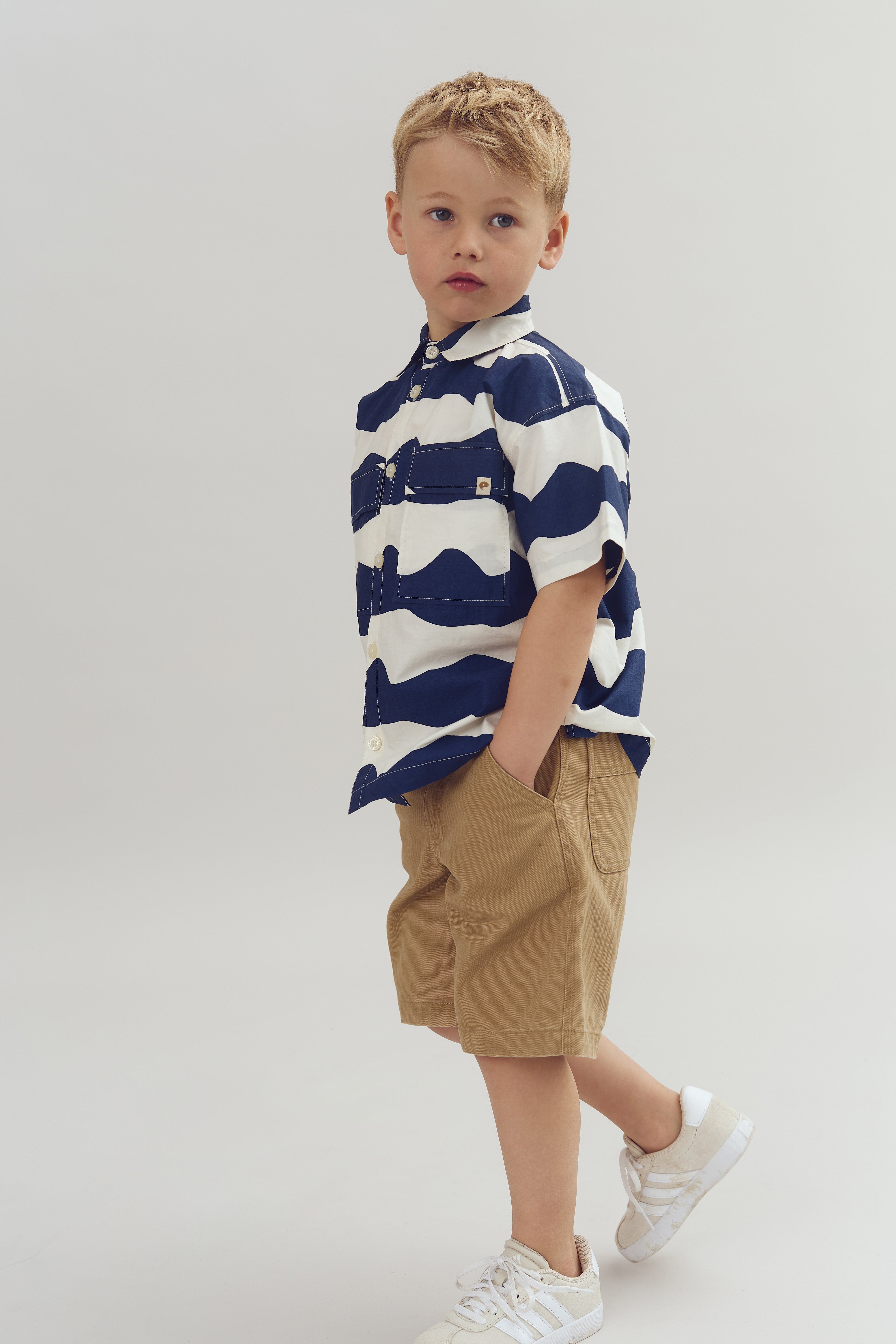 Een jong kind draagt een gestreept overhemd en beige korte broek, staand met handen in zakken. De achtergrond is een eenvoudige, lichte kleur.