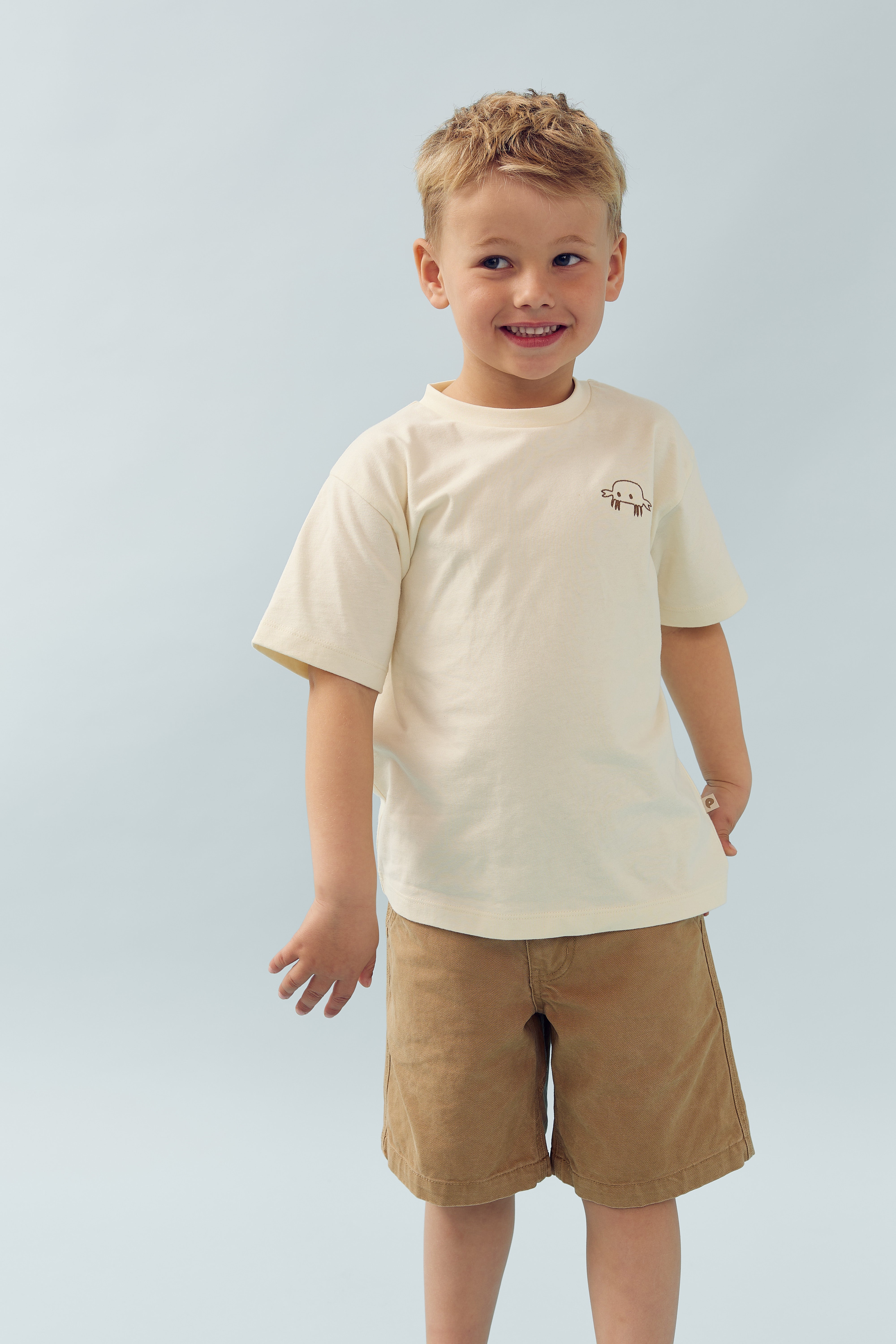 Een jong kind draagt een beige T-shirt met een tekening van een kwal en bruine korte broek, glimlachend tegen een lichte achtergrond.