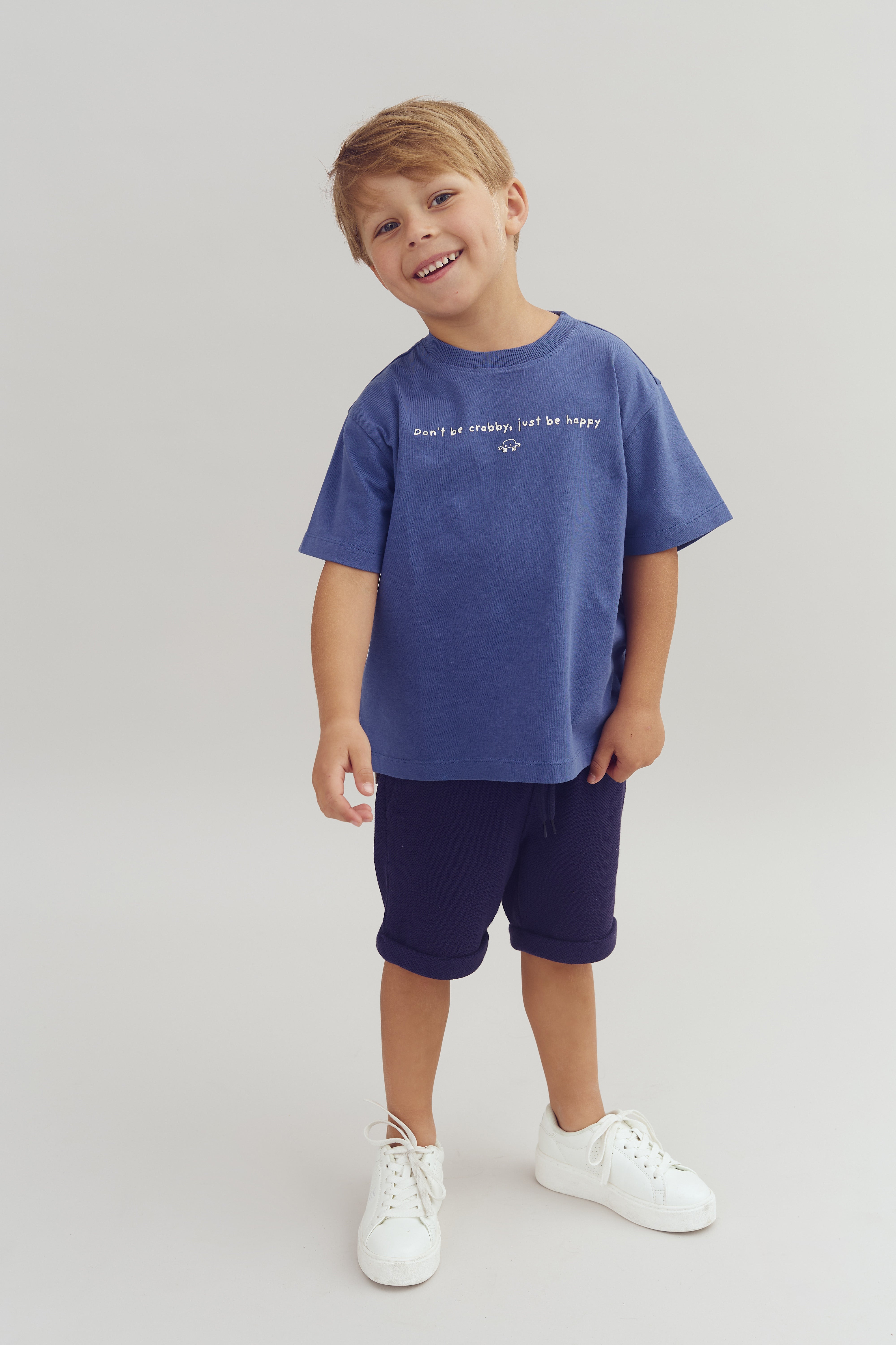 Een kind draagt een blauw T-shirt met de tekst "Don't be crabby, just be happy", lacht en staat tegen een neutrale achtergrond, gekleed in korte broek en witte sneakers.